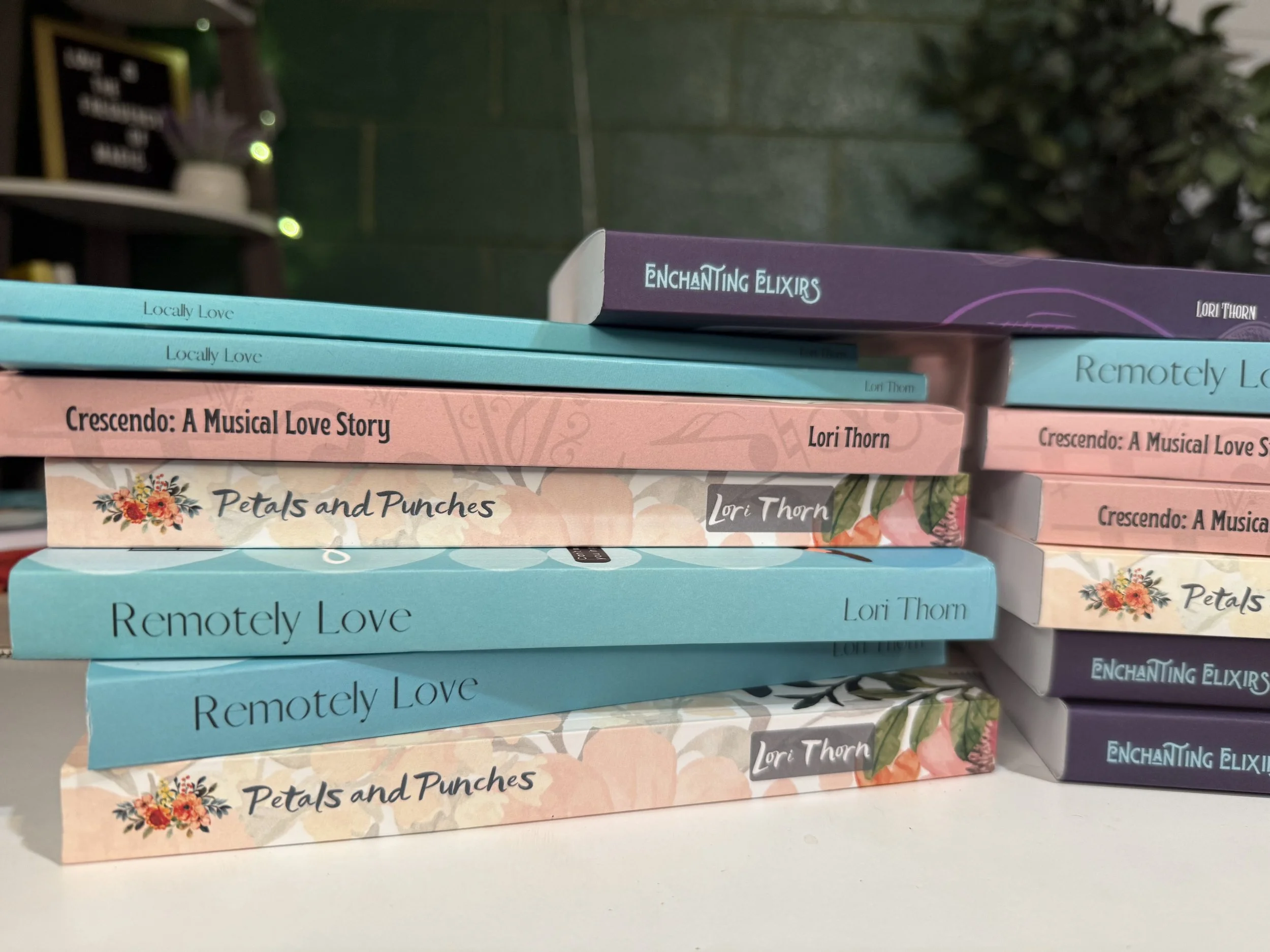 Stacked books with titles including 'Locally Love,' 'Crescendo: A Musical Love Story,' 'Petals and Punches,' 'Remotely Love,' and 'Enchanted Elixirs' by Lori Thorn, arranged on a table with a blurred background.