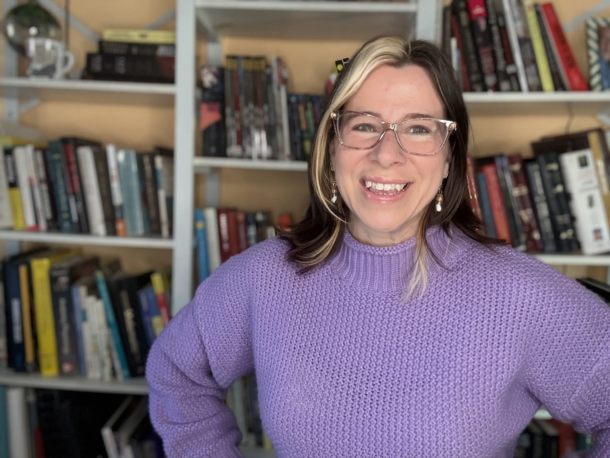 A smiling woman with glasses and earrings wearing a purple sweater in front of a bookshelf filled with books.