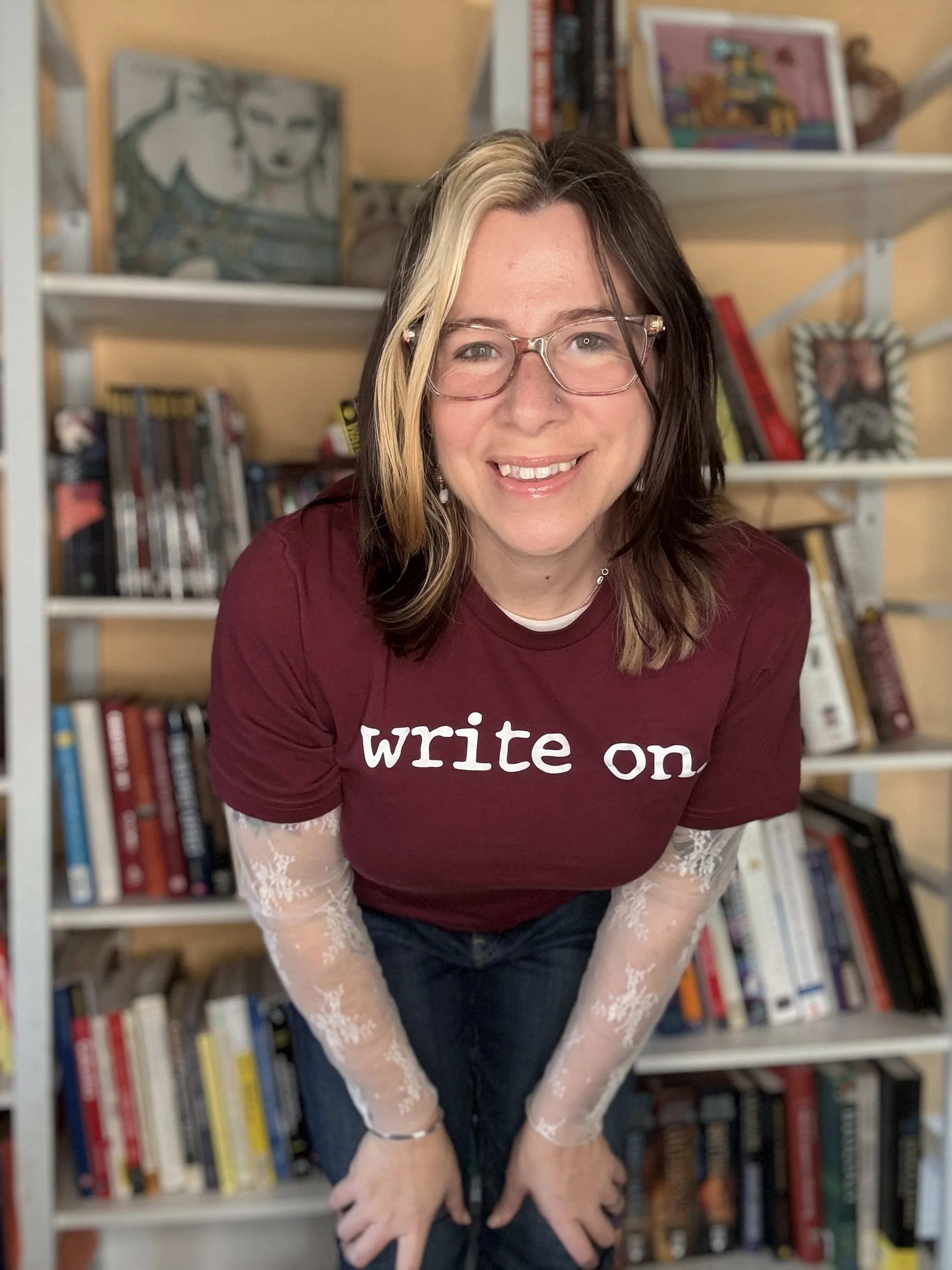 A woman with shoulder length hair, glasses, and a nose piercing, smiling and leaning forward, standing in front of a bookshelf with books and artwork.
