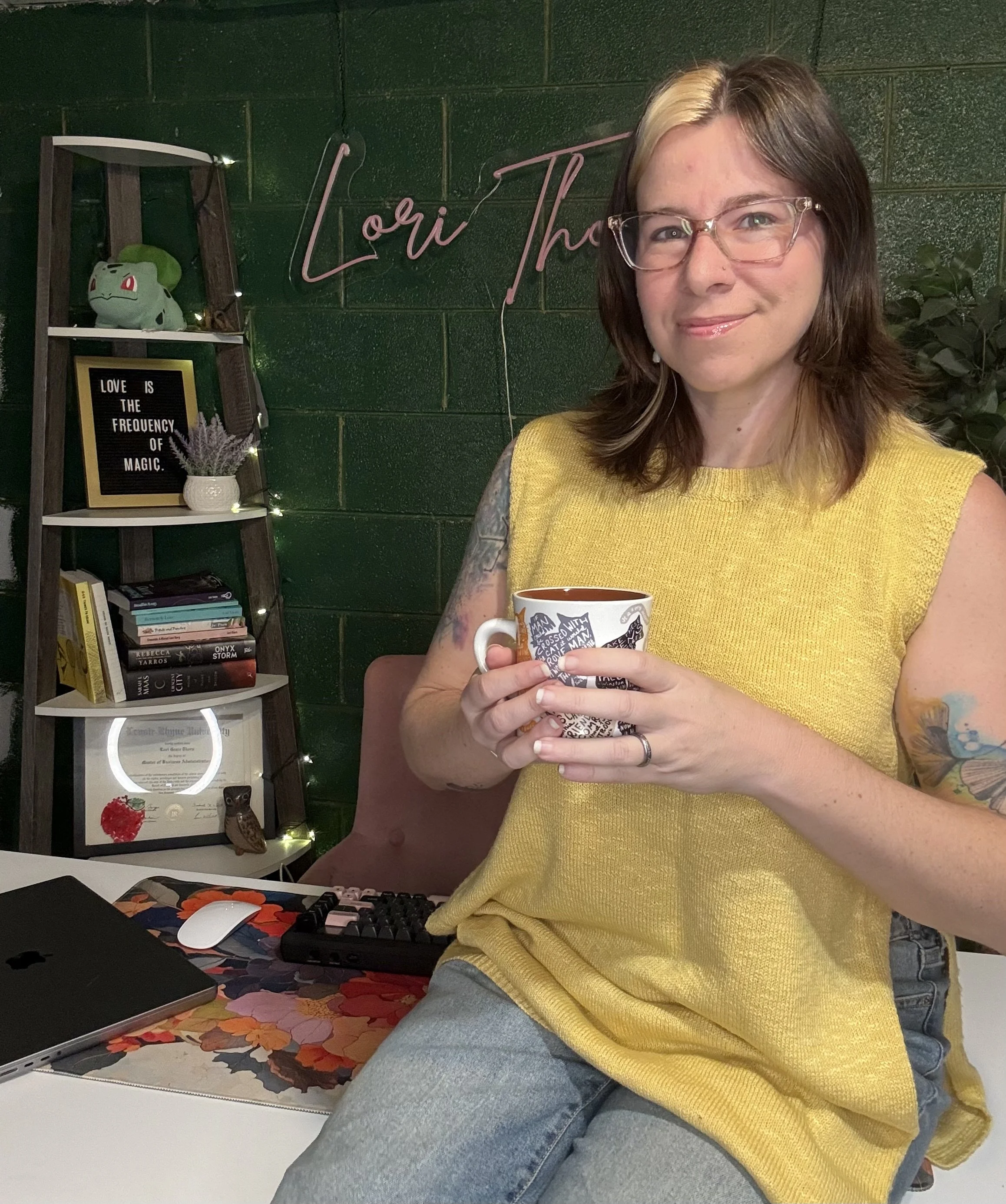 A woman with shoulder-length dark hair, a blonde streak, and glasses, wearing a yellow sleeveless top, holding a mug, sitting in a room decorated with books, a small plant, and string lights, with a green brick wall and a neon sign that says 'Lori Th...'