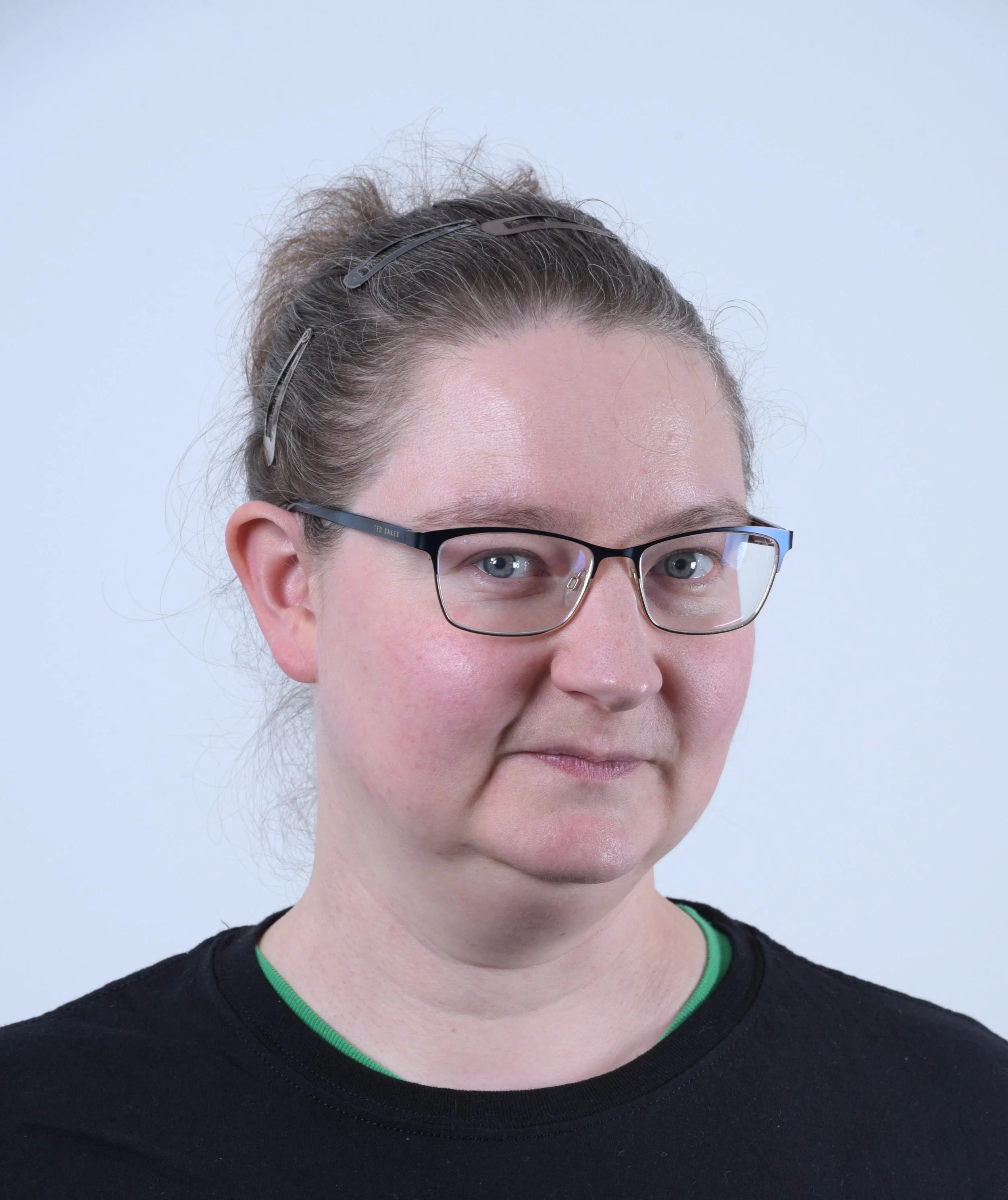 A woman with glasses and hair clips, wearing a black shirt with a green collar, looking at the camera against a plain white background.