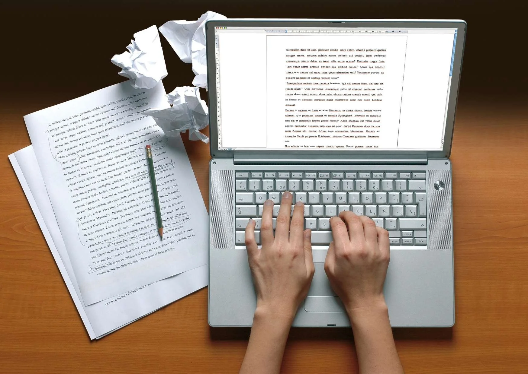 A person typing on a silver laptop with sheets of paper and crumpled paper around it on a wooden desk.