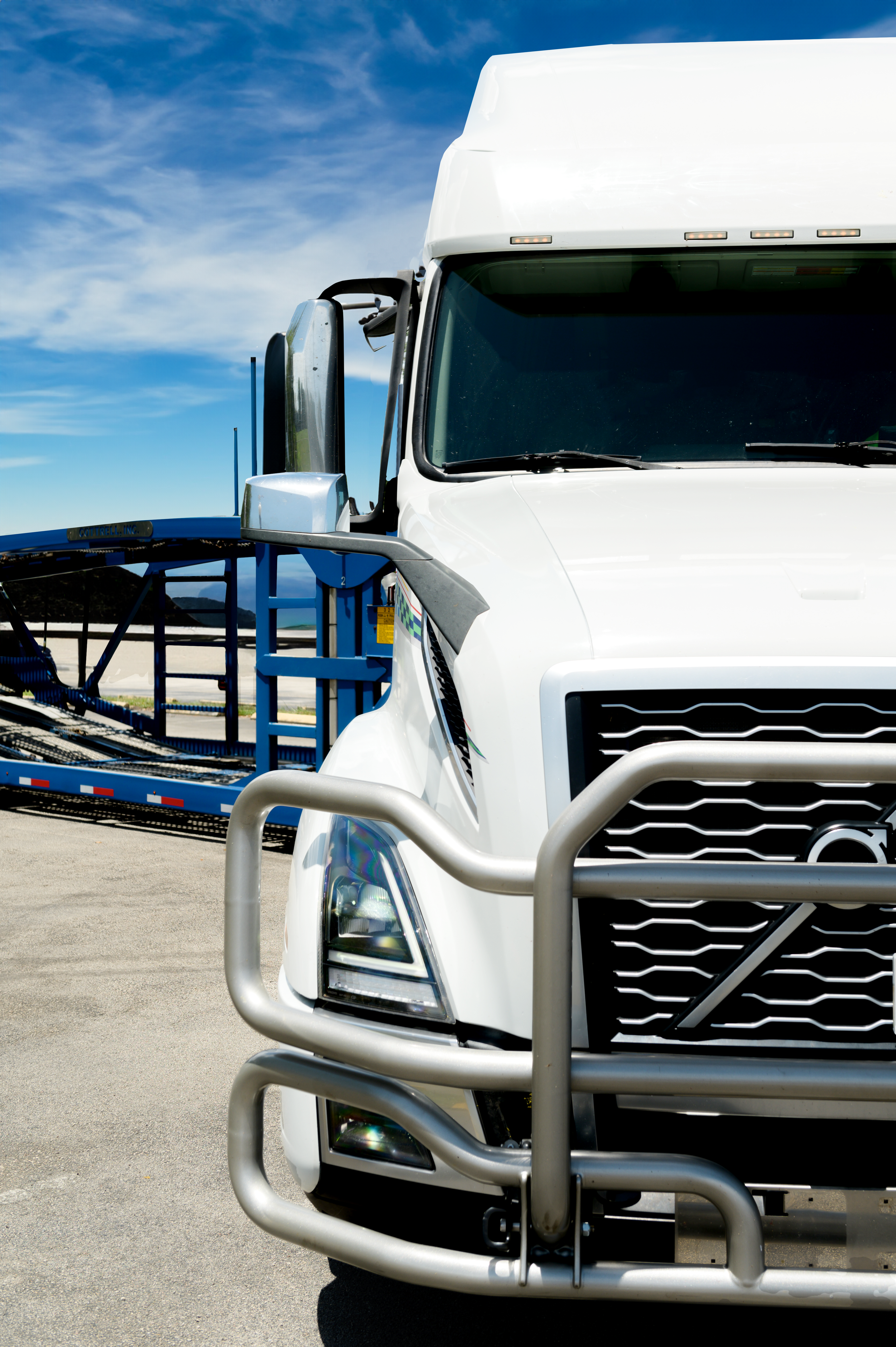 Close-up of a white semi-truck with a protective front bumper, parked near a blue car carrier trailer on a clear day.