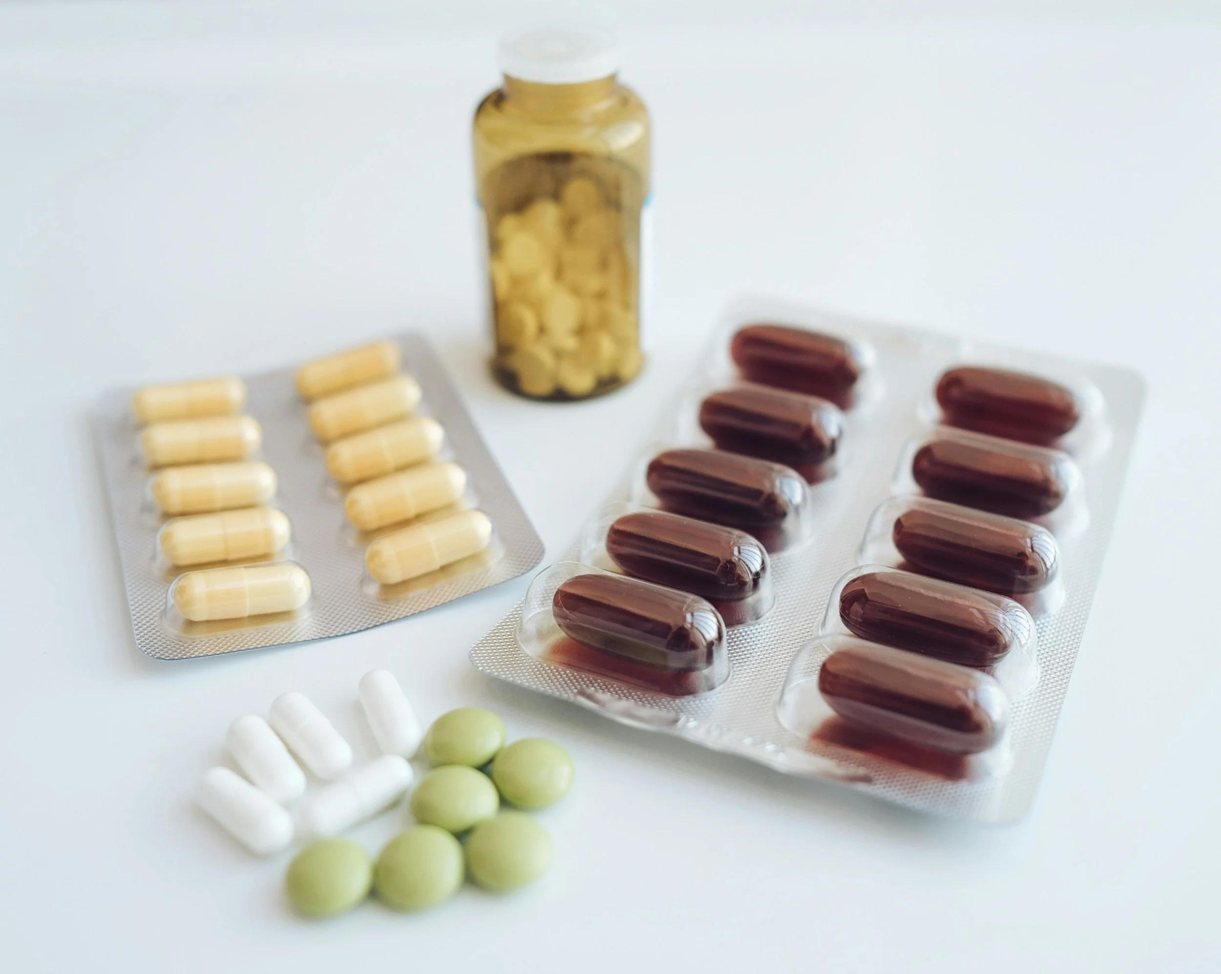 Assorted pills and capsules in blister packs, including white, green, beige, and brown capsules and tablets, with a small vial of yellow pills in the background.