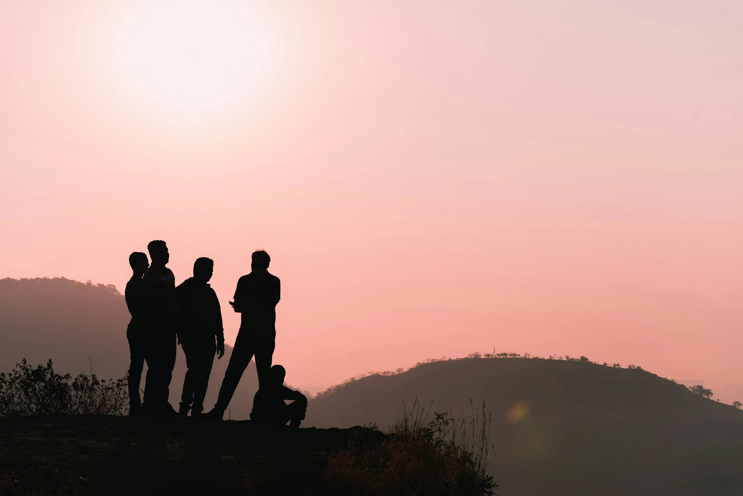 Silhouettes of five people standing on a hilltop during sunset, with a soft pink sky and rolling hills in the background.