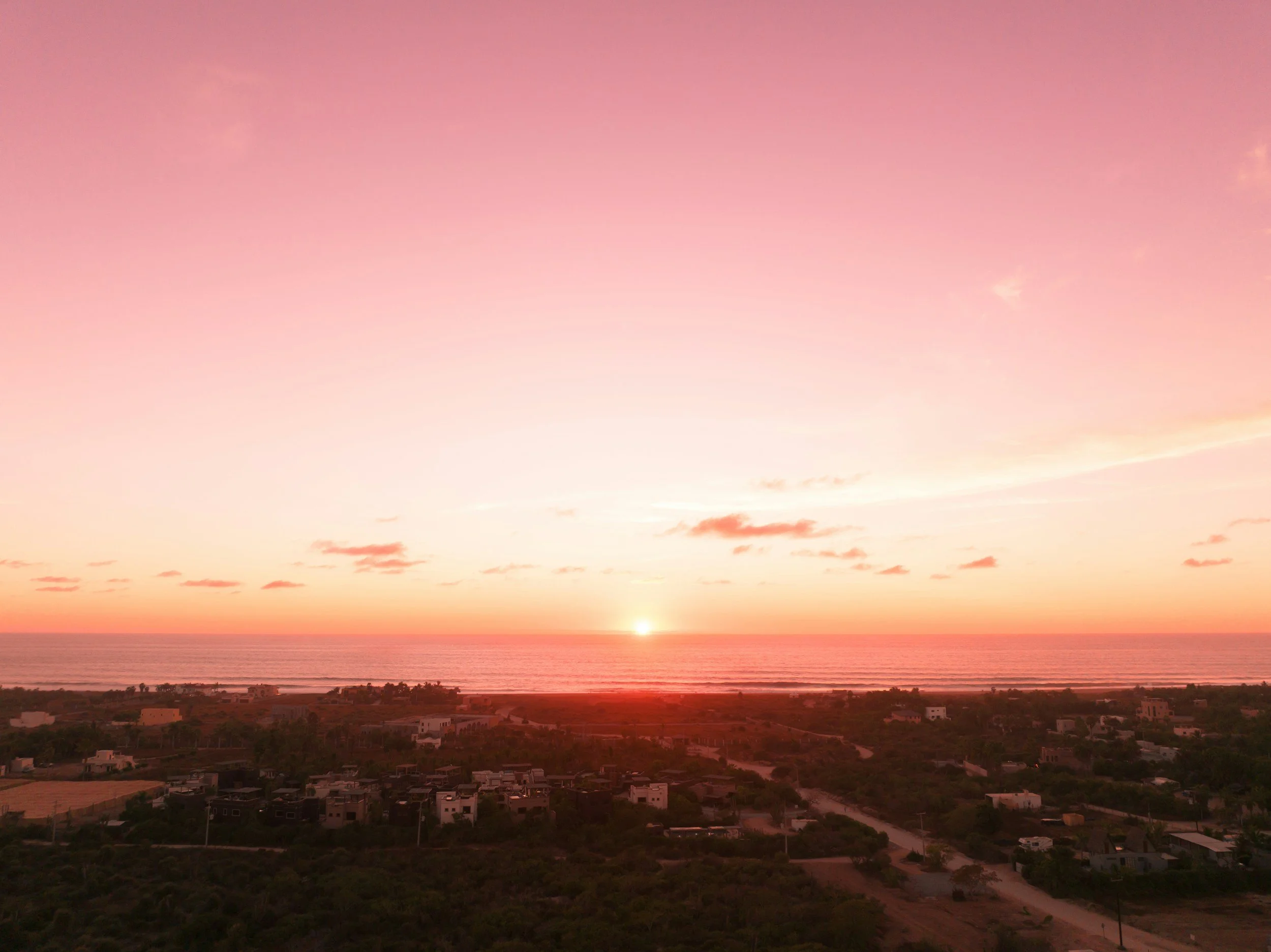 Sunset over the ocean with a pink and orange sky and a few scattered clouds, viewed from a landscape with houses and trees in the foreground.