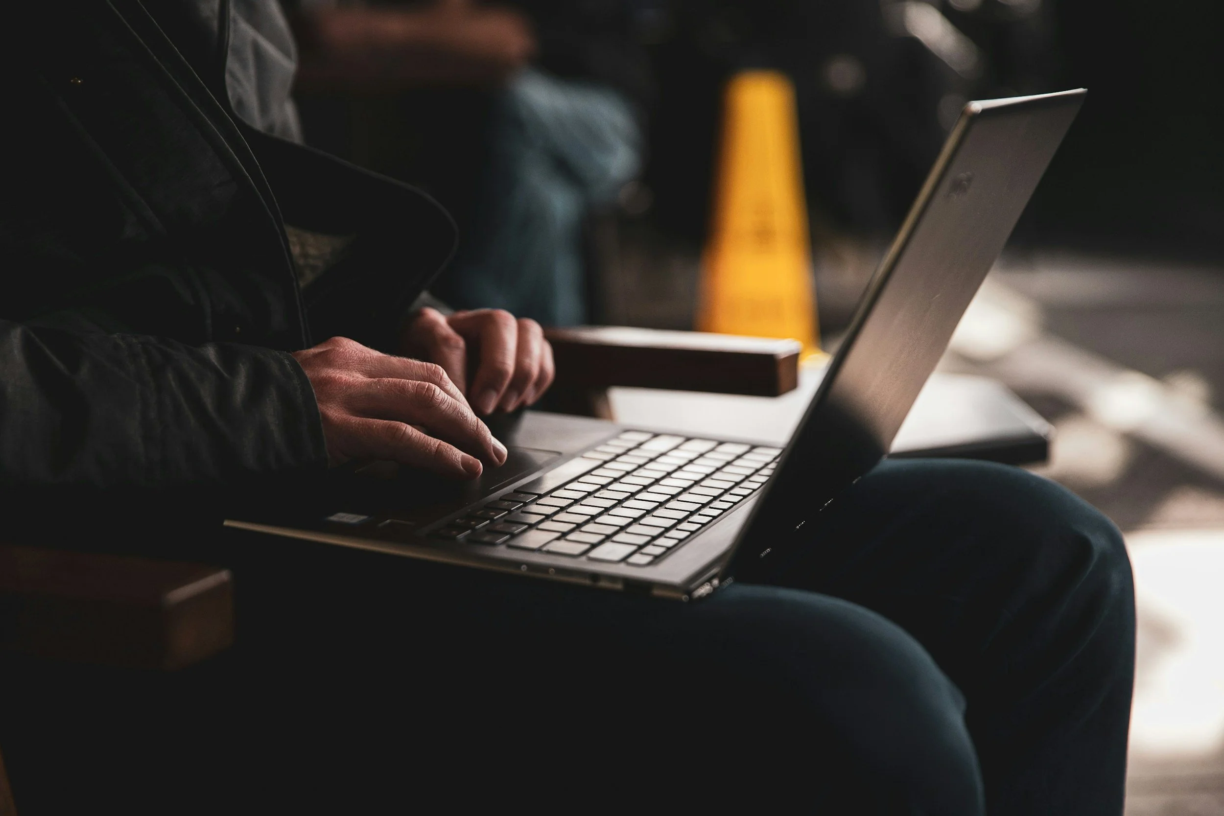 Person using a laptop at a table, with other people in the background on a dark, cozy setting.