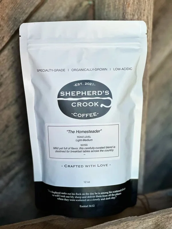 White coffee bag from Shepherd's Crook Coffee with black and white label, labeled 'The Homesteader' roast, 12 oz, featuring a circular logo, and describing the coffee as light-medium roast with mild yet full flavor.