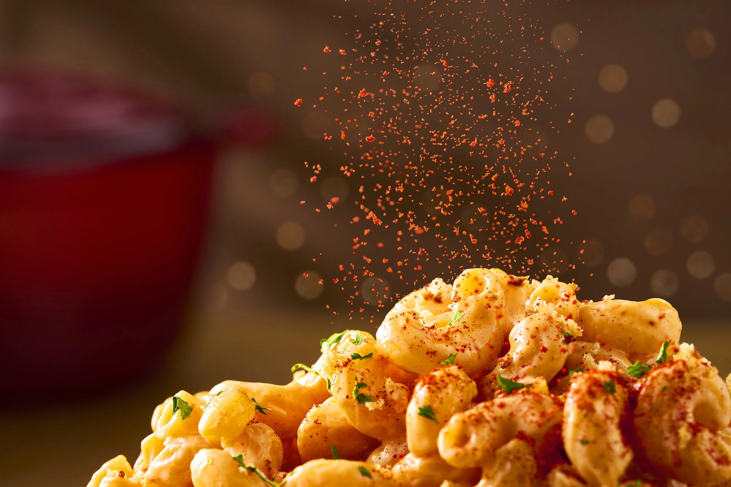 Close-up of macaroni and cheese sprinkled with paprika and chopped herbs, with more paprika being sprinkled from above.