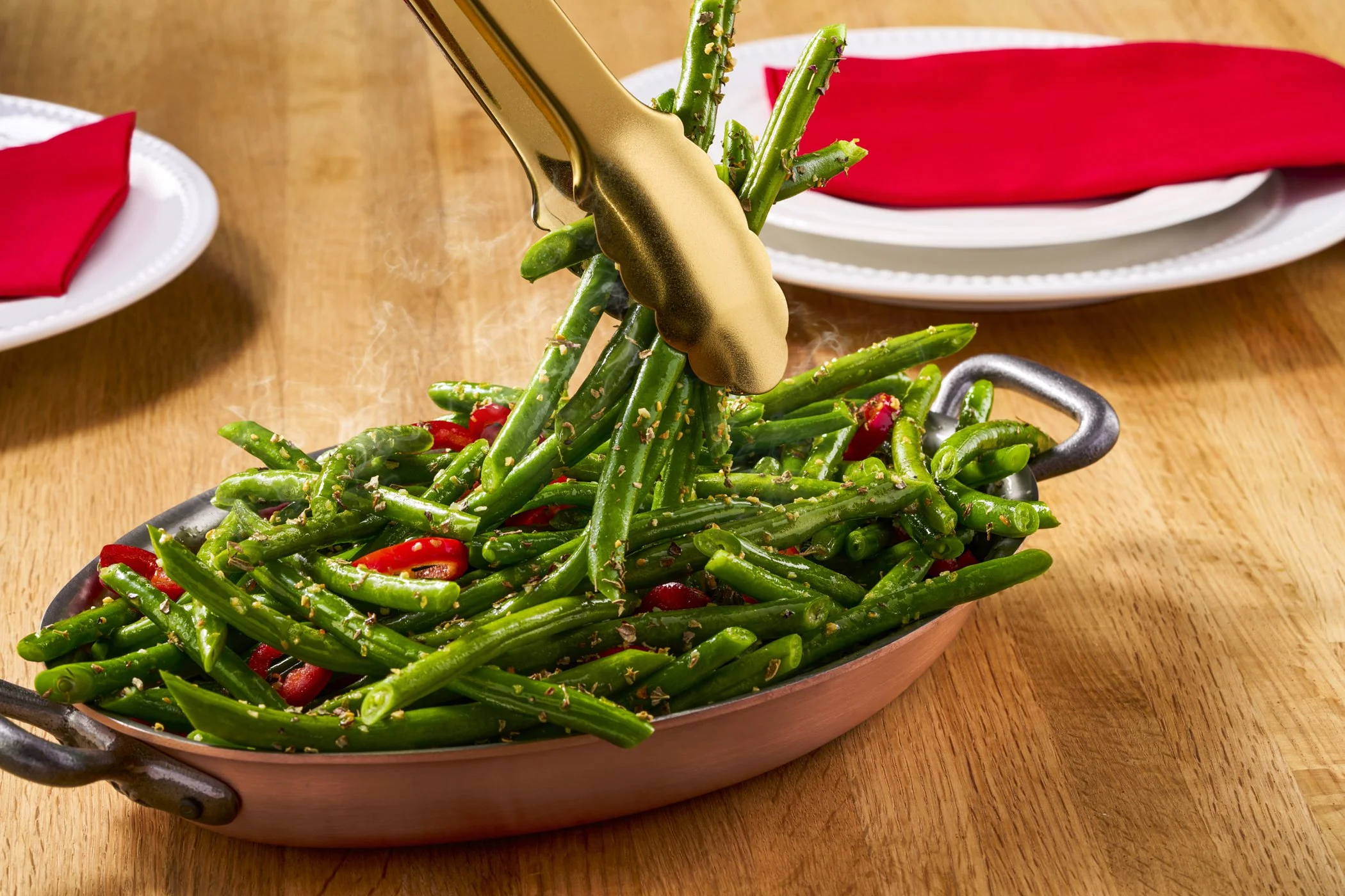Tongs holding fresh green beans and red chili peppers on a serving dish over a wooden table, with plates and red napkins in the background.