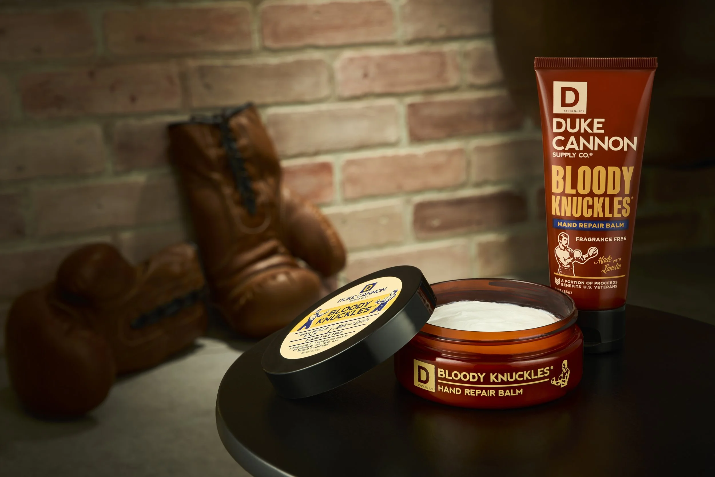A container of Duke Cannon Bloody Knuckles hand repair balm open on a black table, with a tube of the same balm standing upright next to it, and a pair of brown leather work boots in the background against a brick wall.