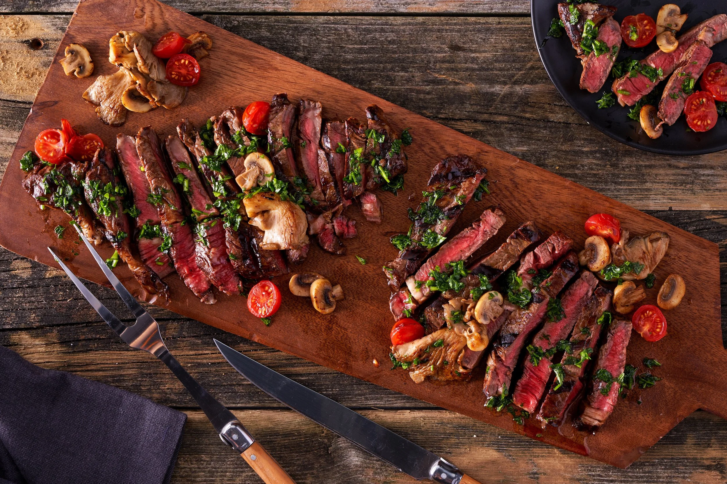 Sliced grilled steak topped with chopped parsley and cherry tomatoes on a wooden serving board, with mushrooms and more cherry tomatoes.
