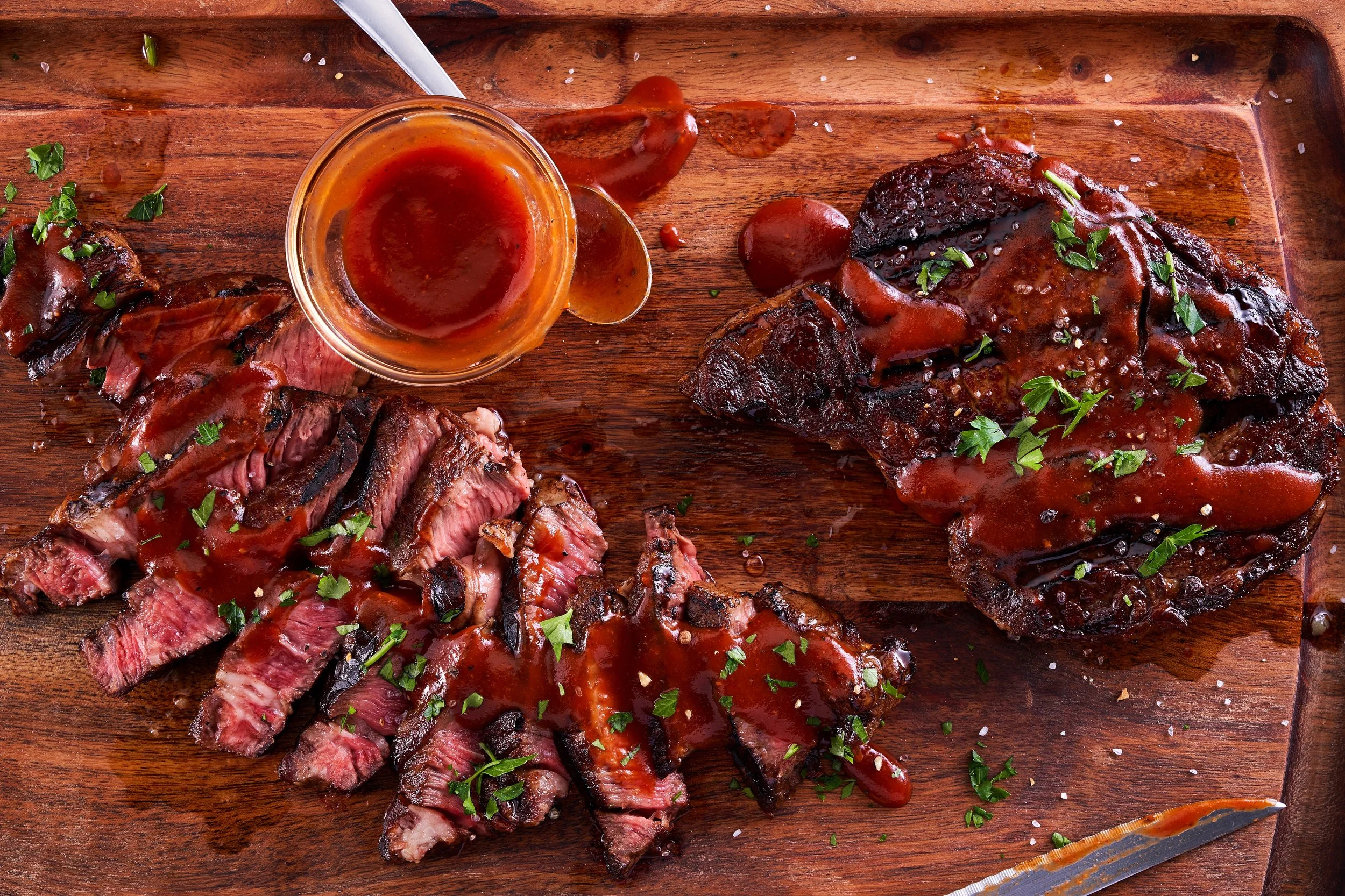 Sliced steak and a full piece of beef brisket on a wooden cutting board with barbecue sauce and parsley garnish.