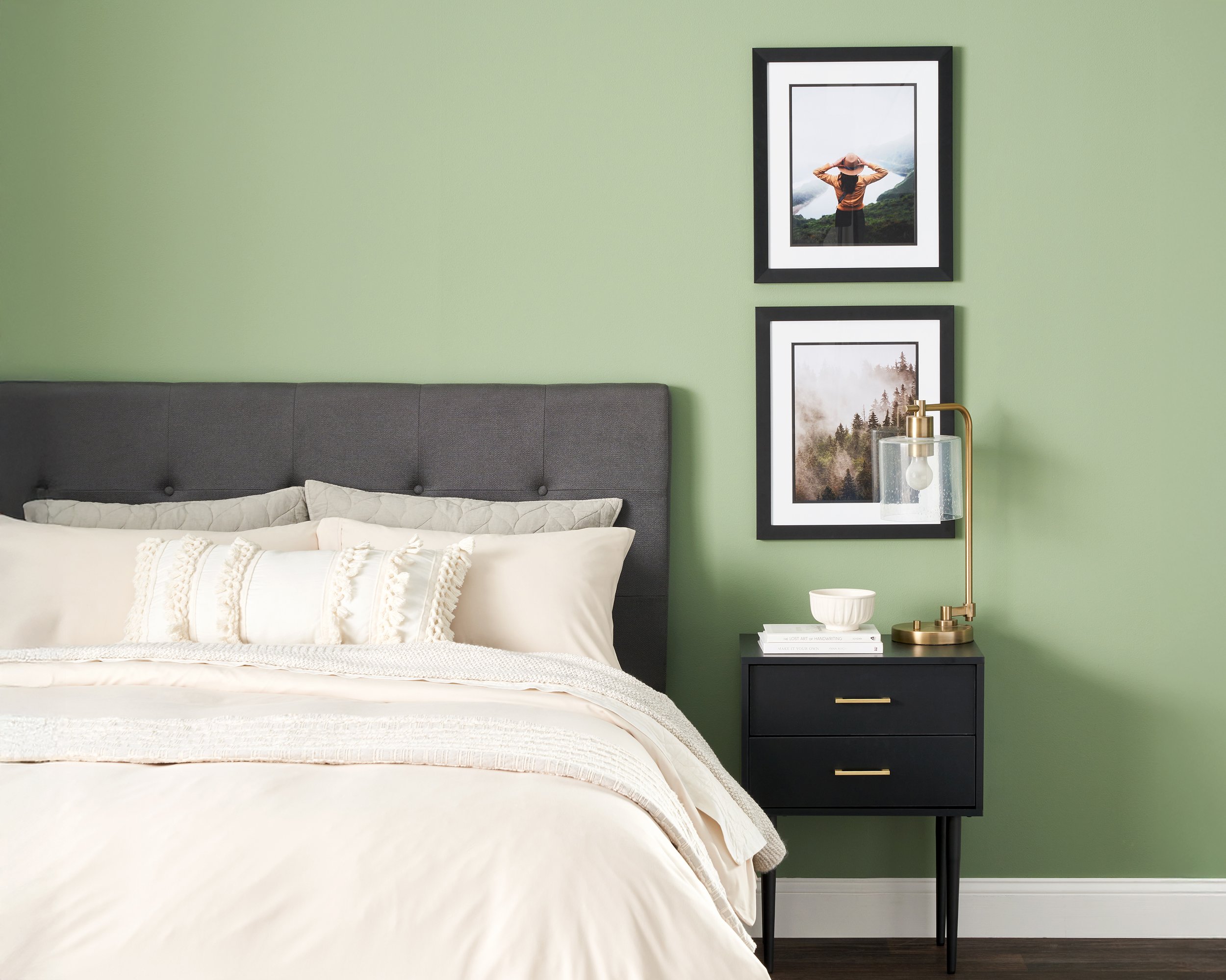 Bedroom with a gray headboard, white bedding, and a black nightstand with gold accents, a gold and glass table lamp, and framed landscape photos on a green wall.