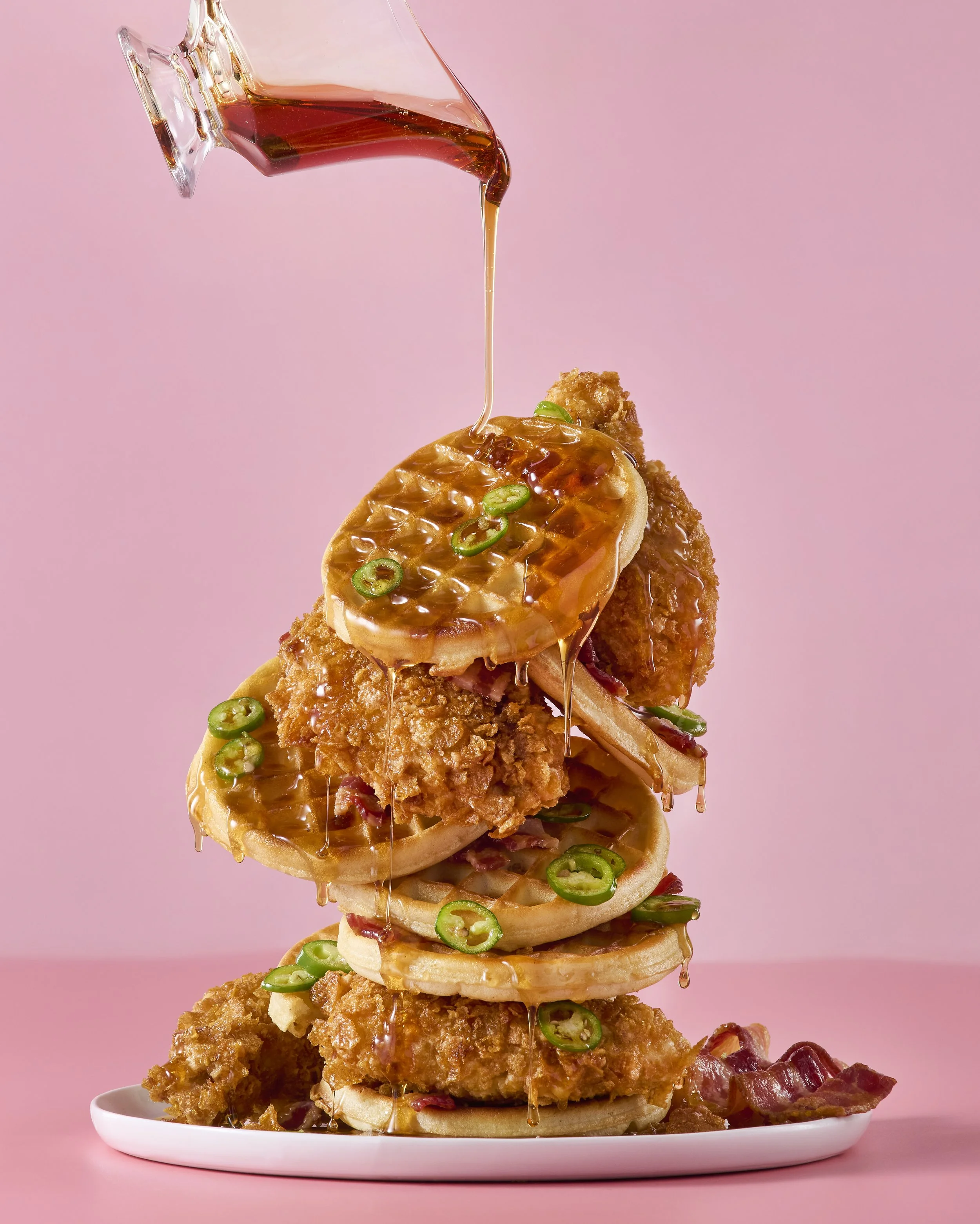 A tower of fried chicken, onion rings, and green pepper slices, topped with a slice of pineapple and sauce being poured from a glass.