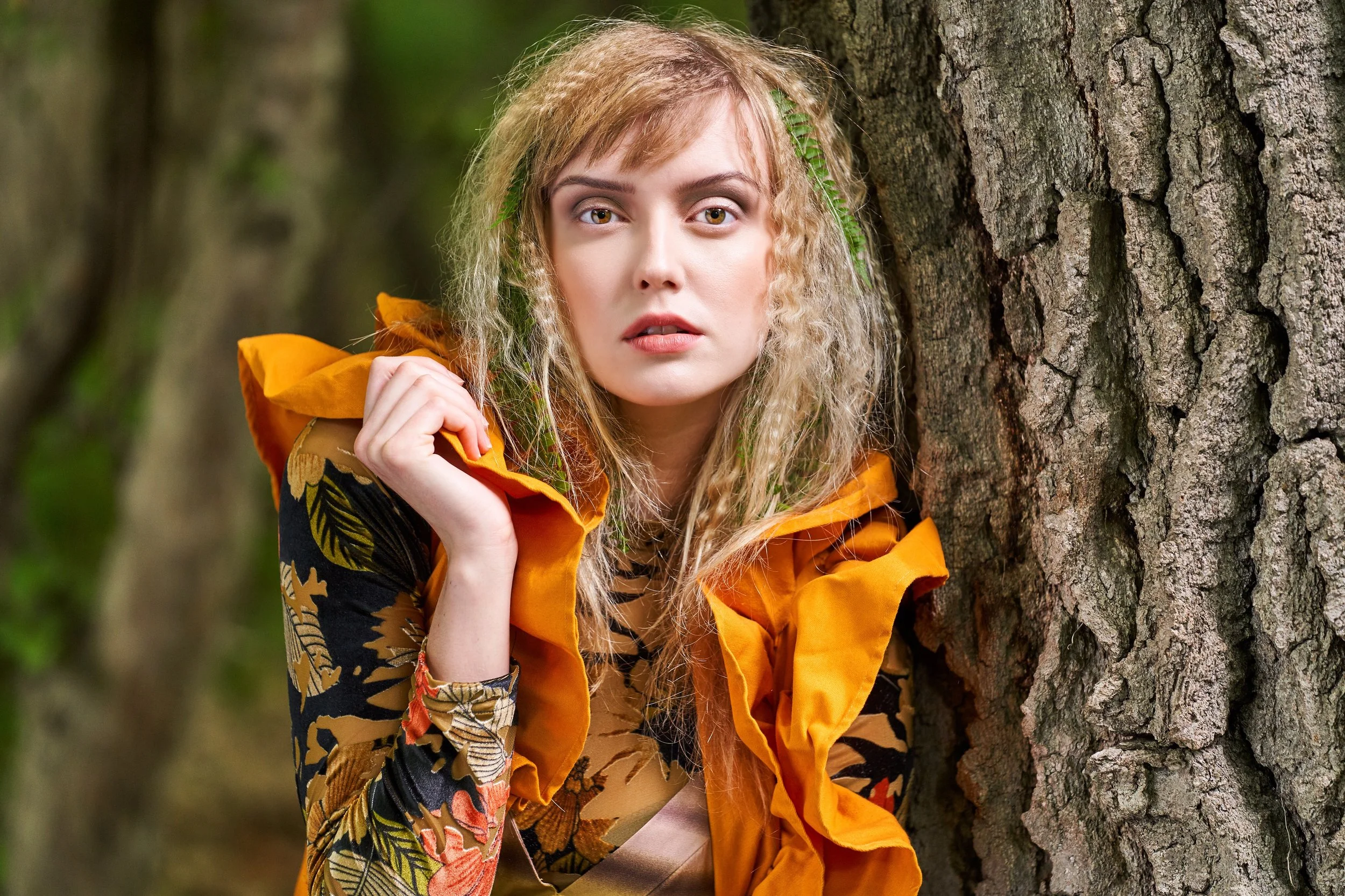 A woman with curly blonde hair and light skin standing next to a large tree trunk in a forest, wearing an orange and floral patterned jacket.