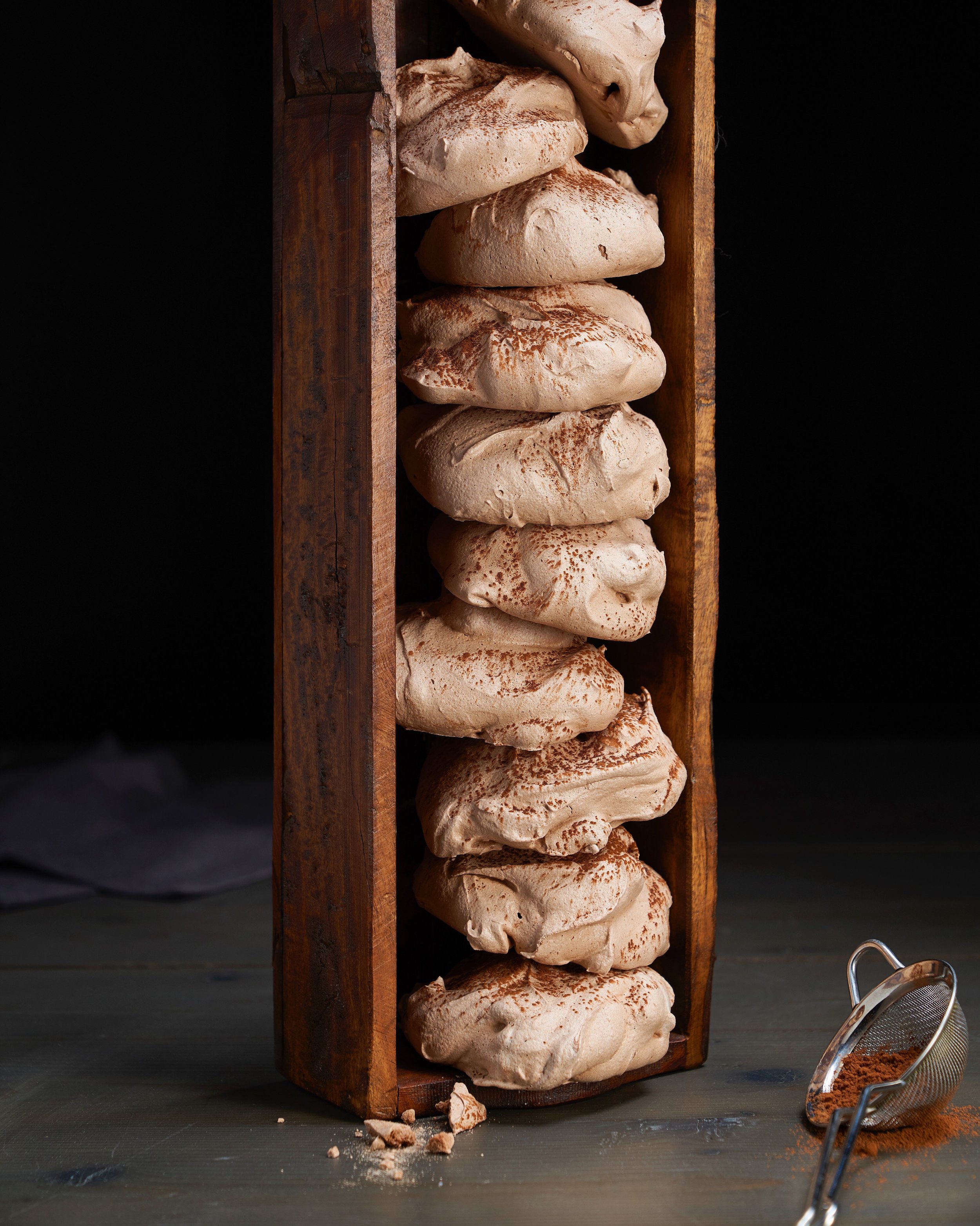 Stacked meringue cookies with cocoa powder in a wooden frame on a dark surface, with a small strainer filled with cocoa powder beside them.