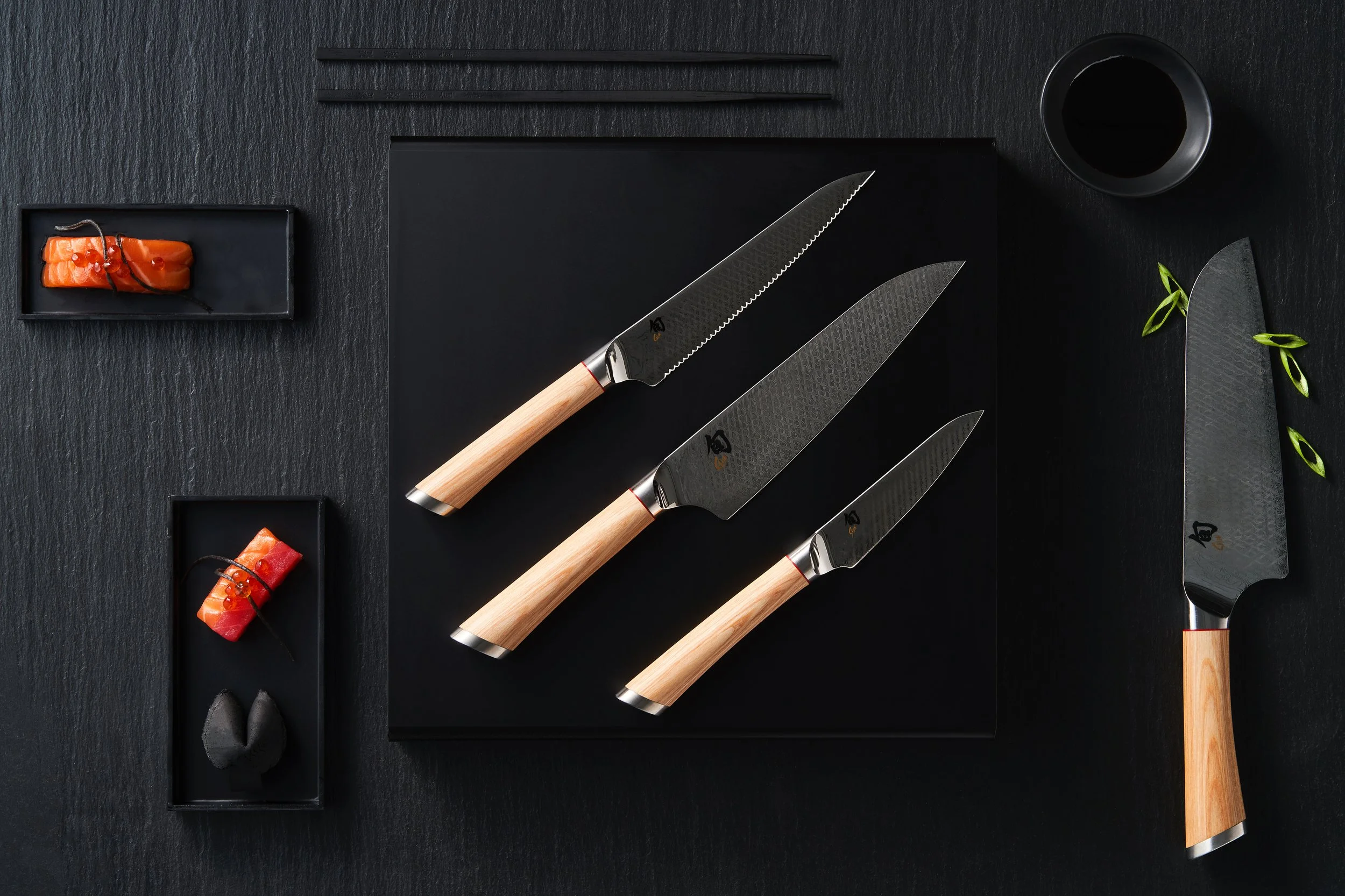 A flat lay of four Japanese-style knives with wooden handles on a black textured surface, accompanied by small rectangular black dishes holding slices of salmon sashimi, black chopsticks, a small black bowl of soy sauce, and a black knife sheath with