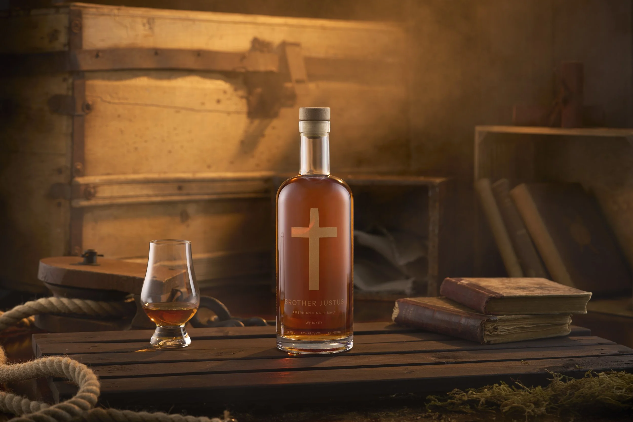 A bottle of Brother Justus American single malt whiskey, a whiskey glass with whiskey, and two old books on a wooden surface in a rustic setting.