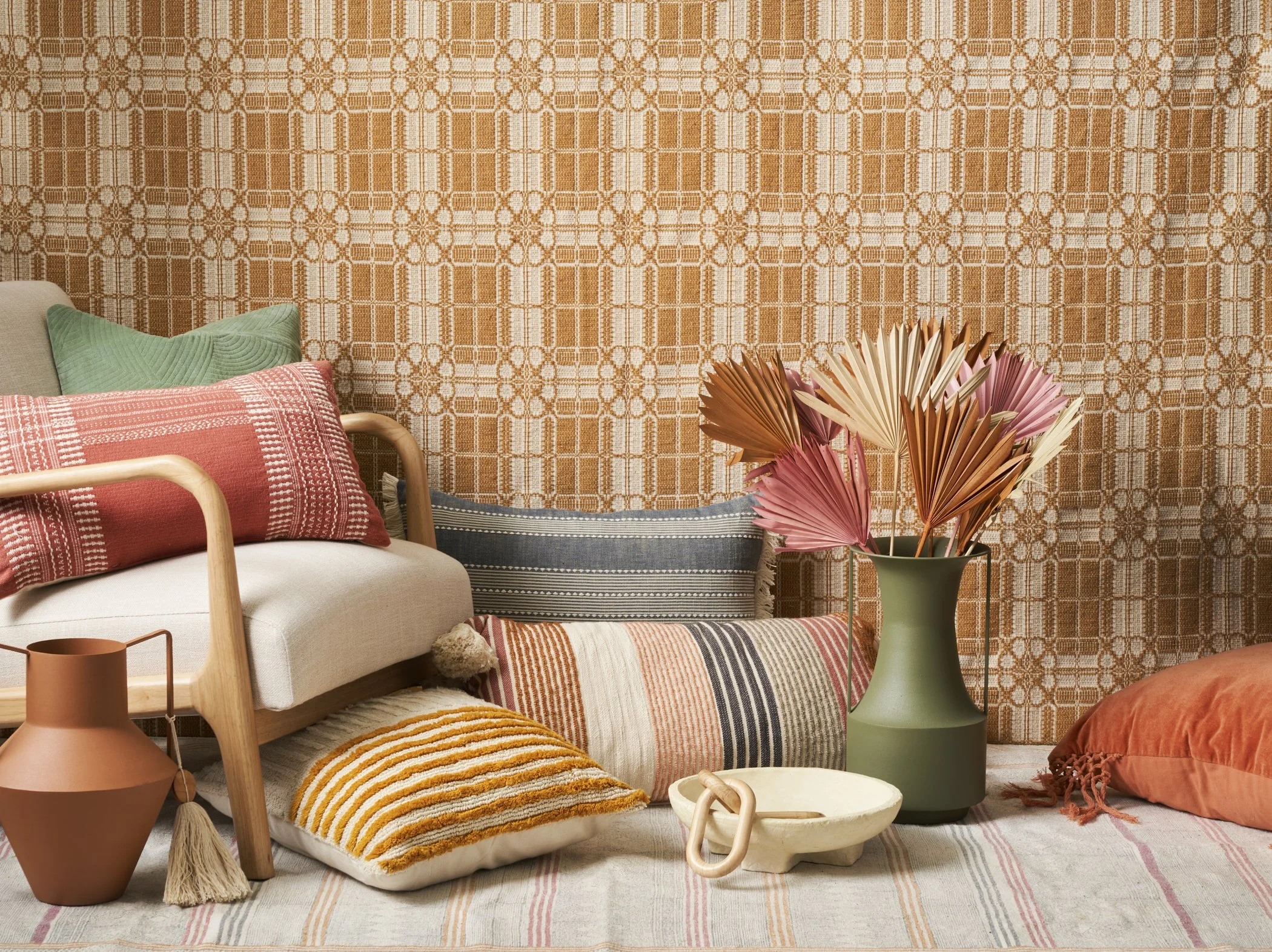 Interior scene with a beige sofa, colorful cushions, a large green vase with dried pink and beige palm leaves, and decorative items on the floor, all set against a patterned orange and white background.