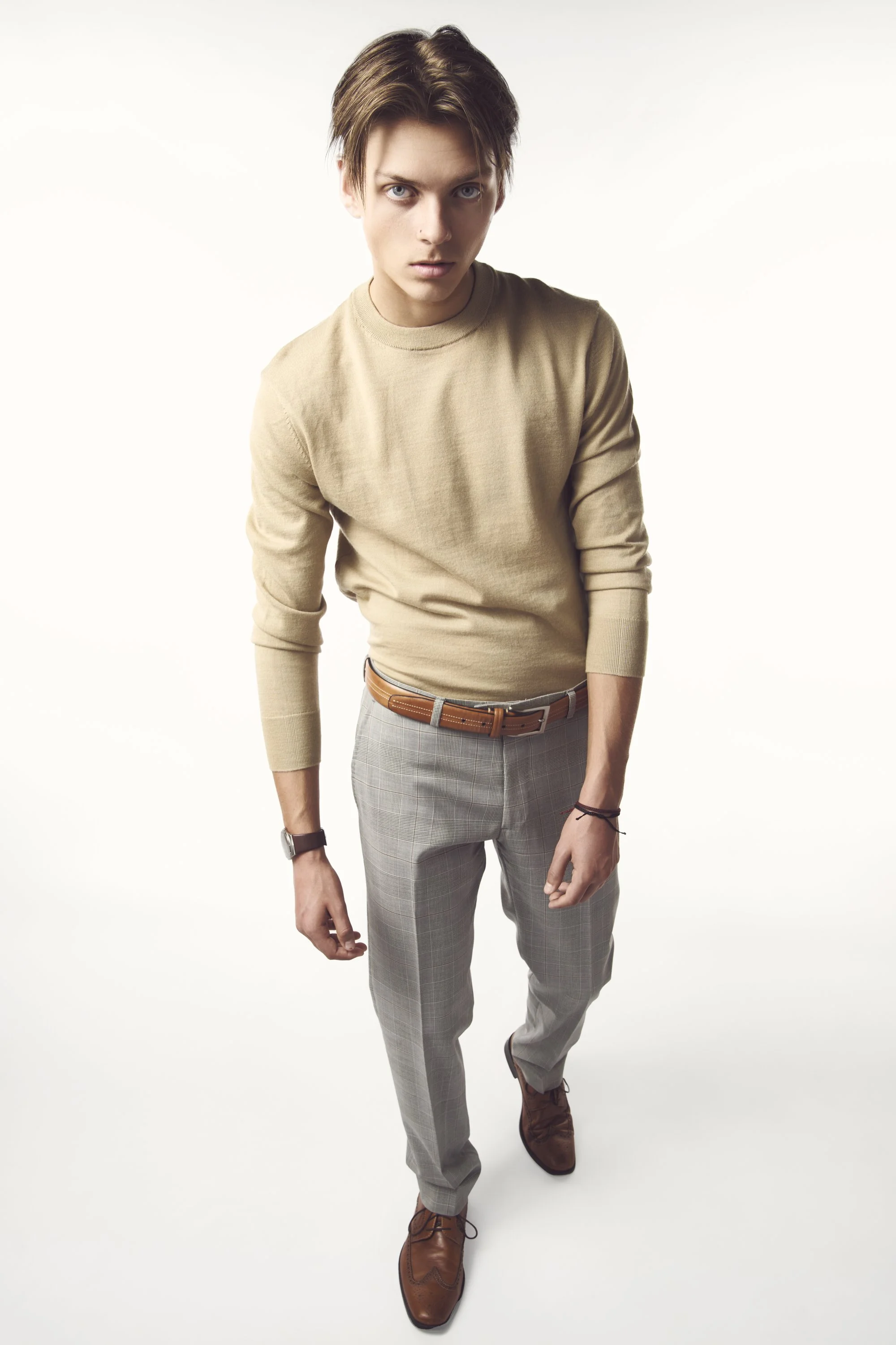 A young man with brown hair, wearing a beige sweater, gray plaid pants, brown shoes, and a black watch, standing against a white background.