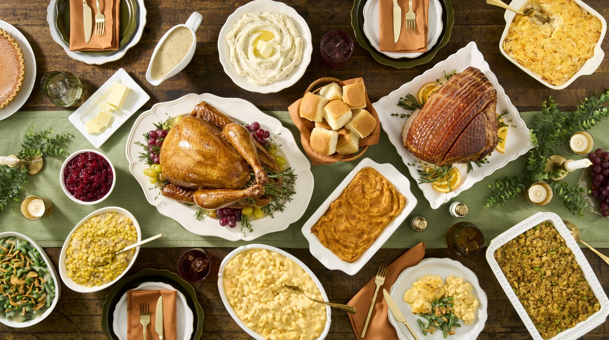 A Thanksgiving feast with a roasted turkey, ham, mashed potatoes, macaroni and cheese, green beans, stuffing, cranberry sauce, bread rolls, and various side dishes on a wooden table decorated with candles, greenery, and orange napkins.