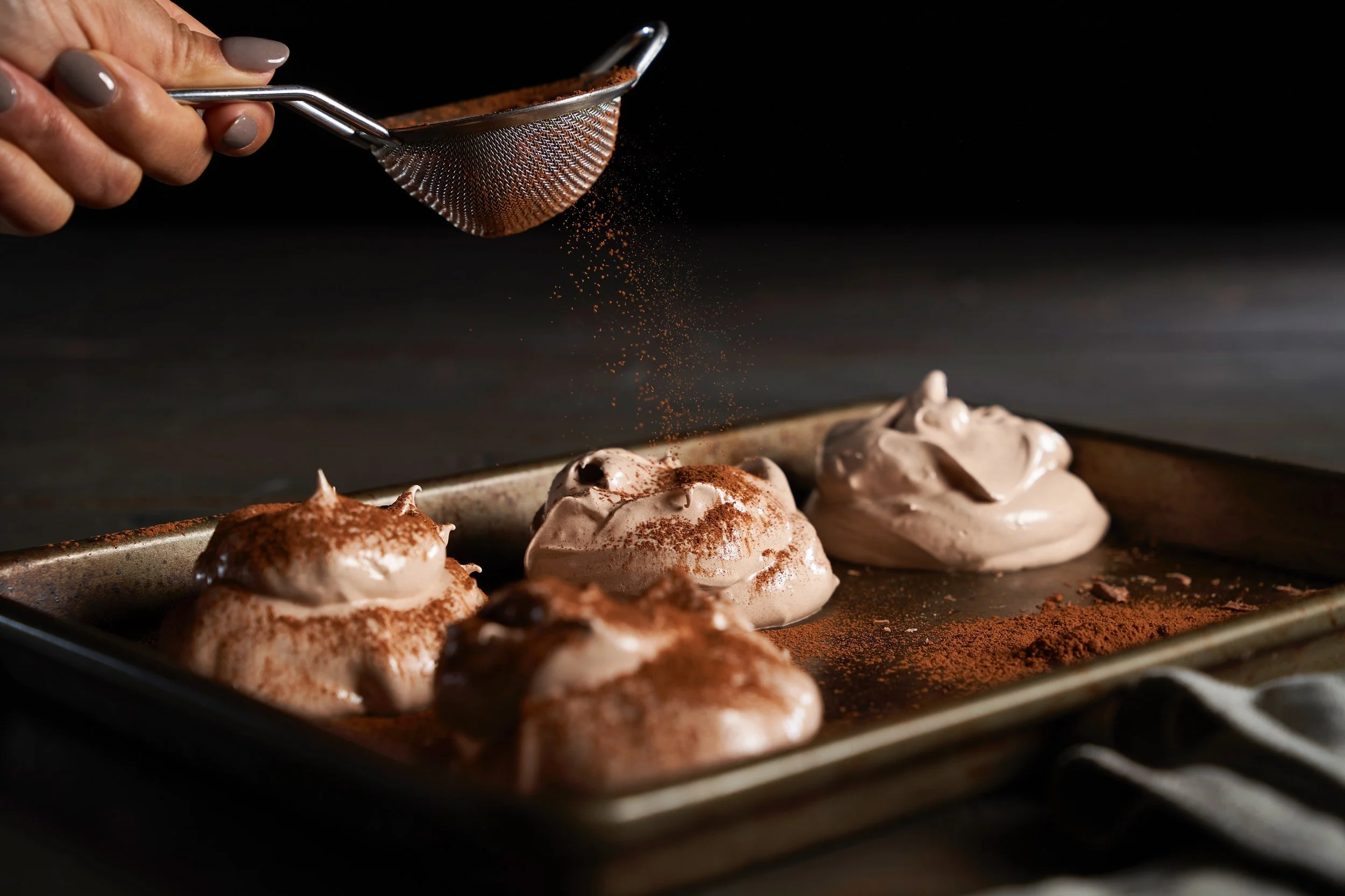 Hand sprinkling cocoa powder over meringue desserts on a baking tray, dark background.