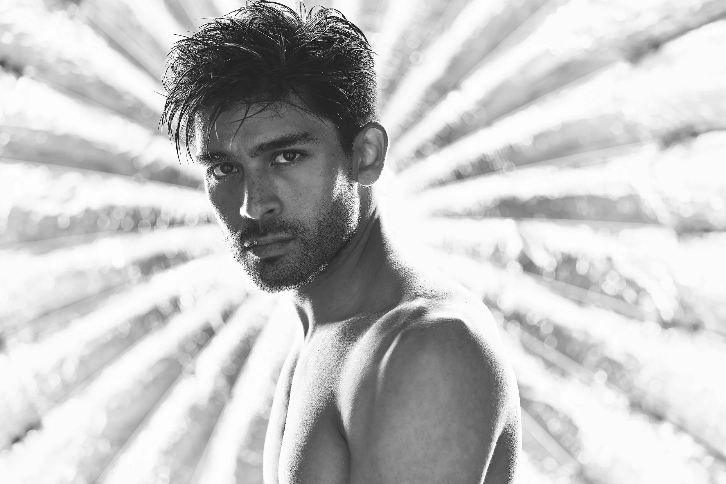 A black and white photo of a shirtless man with dark, tousled hair and facial hair, looking at the camera with a serious expression. The background features blurred, radiating lines.