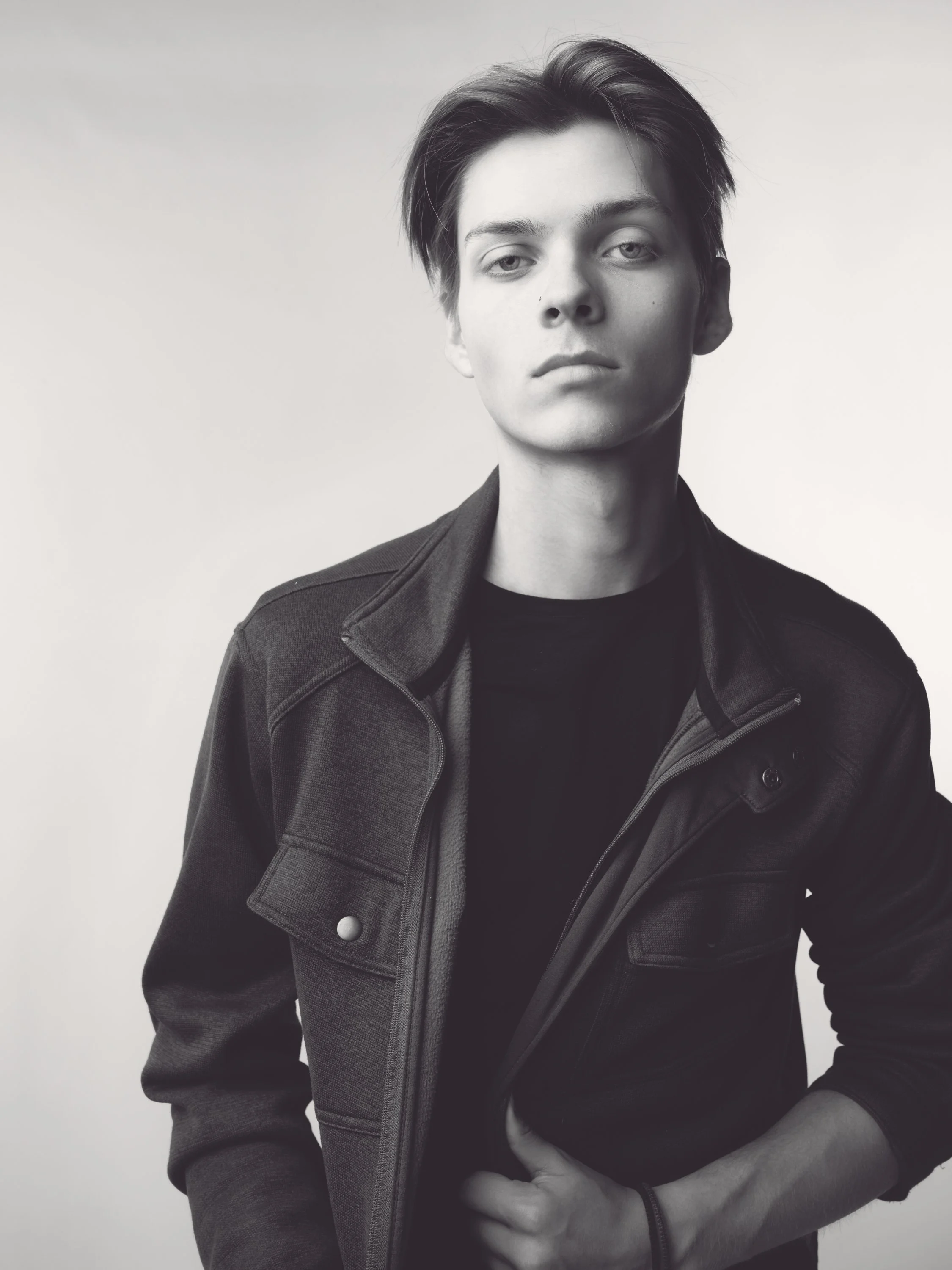 Black and white portrait of a young man with short hair, wearing a jacket over a black shirt, standing against a plain light background.