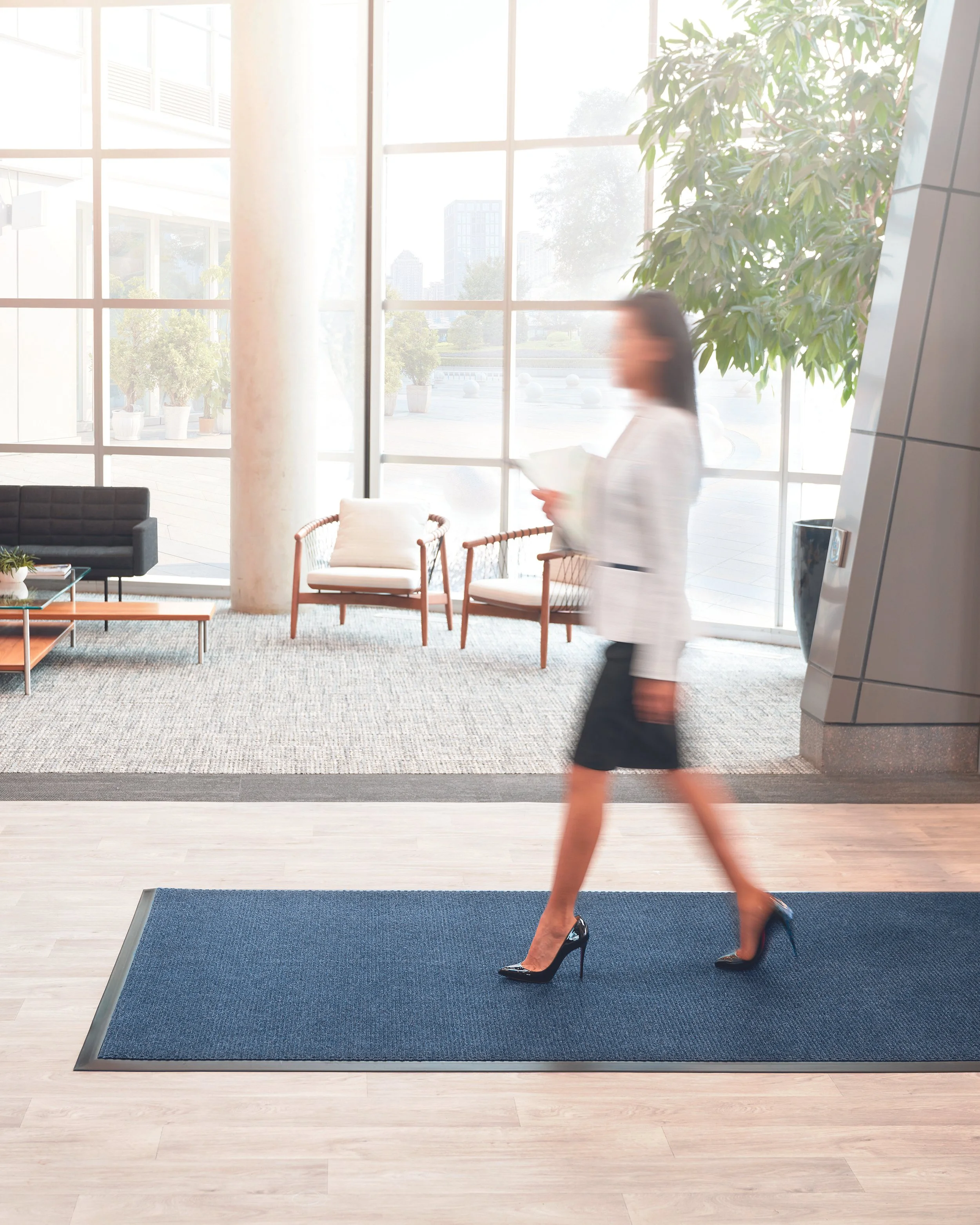 Woman walking through an office lobby, wearing black high heels and a white blouse, with large windows letting in natural light and potted plants outside.