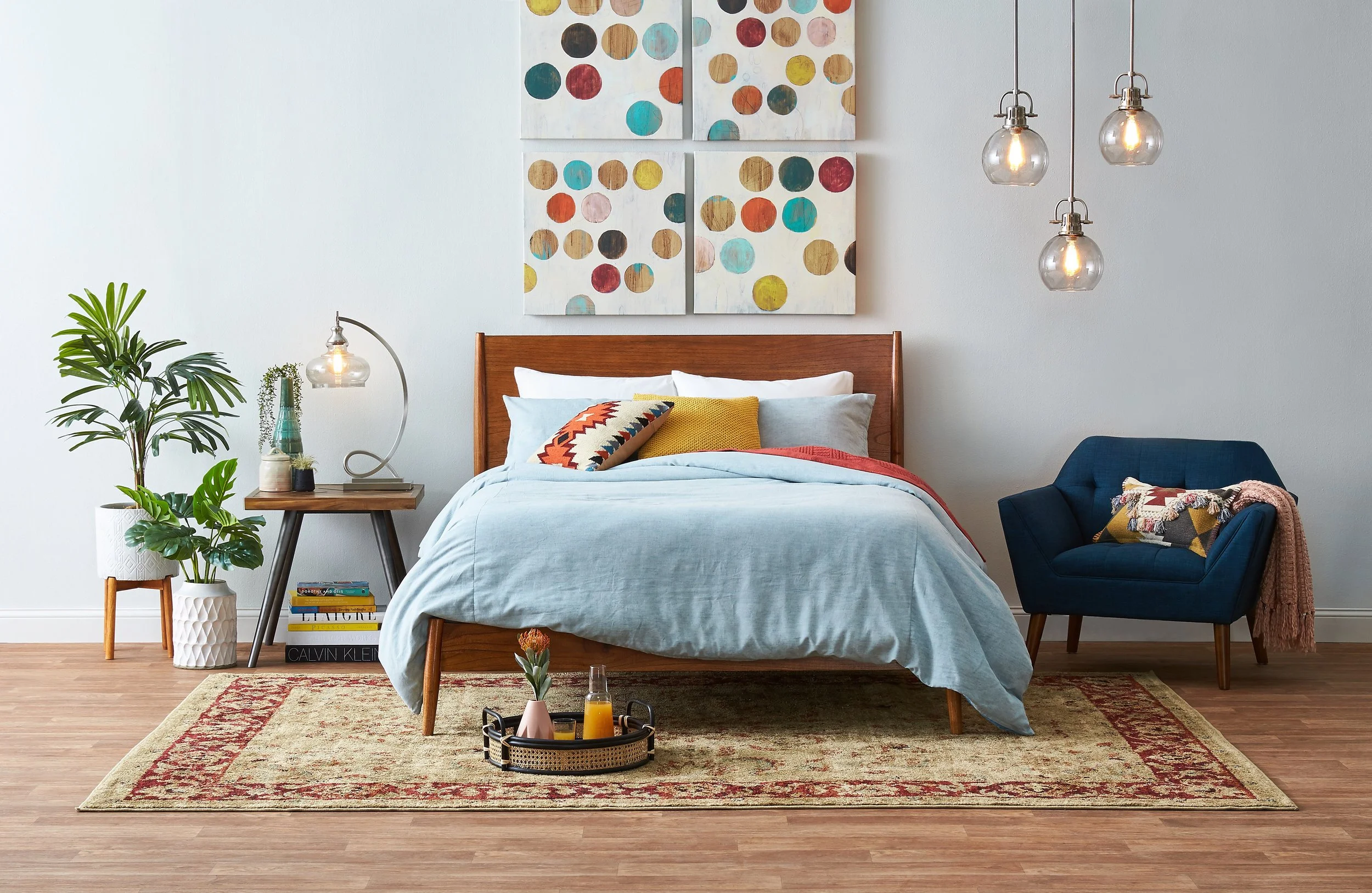 A bedroom with a wooden bed frame, light blue bedding, decorative pillows, a side table with a lamp, potted plants, a blue armchair with pillows and a throw blanket, hanging pendant lights, wall art, and a patterned rug on a wooden floor.