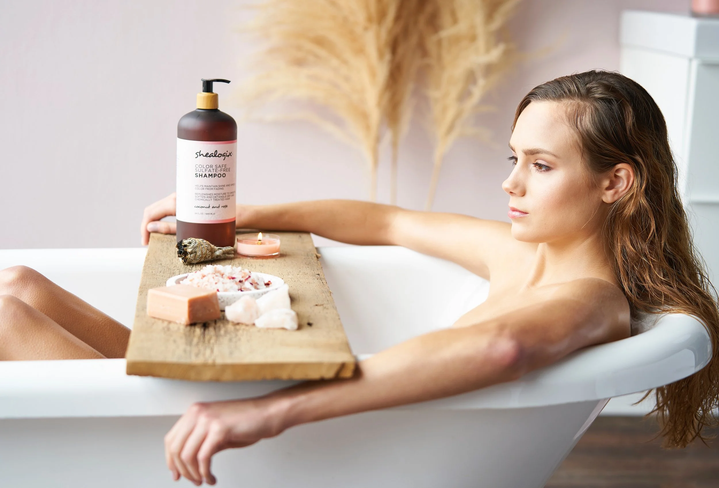 A woman with long, wavy hair relaxing in a bathtub with a wooden tray holding bath products, soap, a lit candle, and a bowl of bath salts.