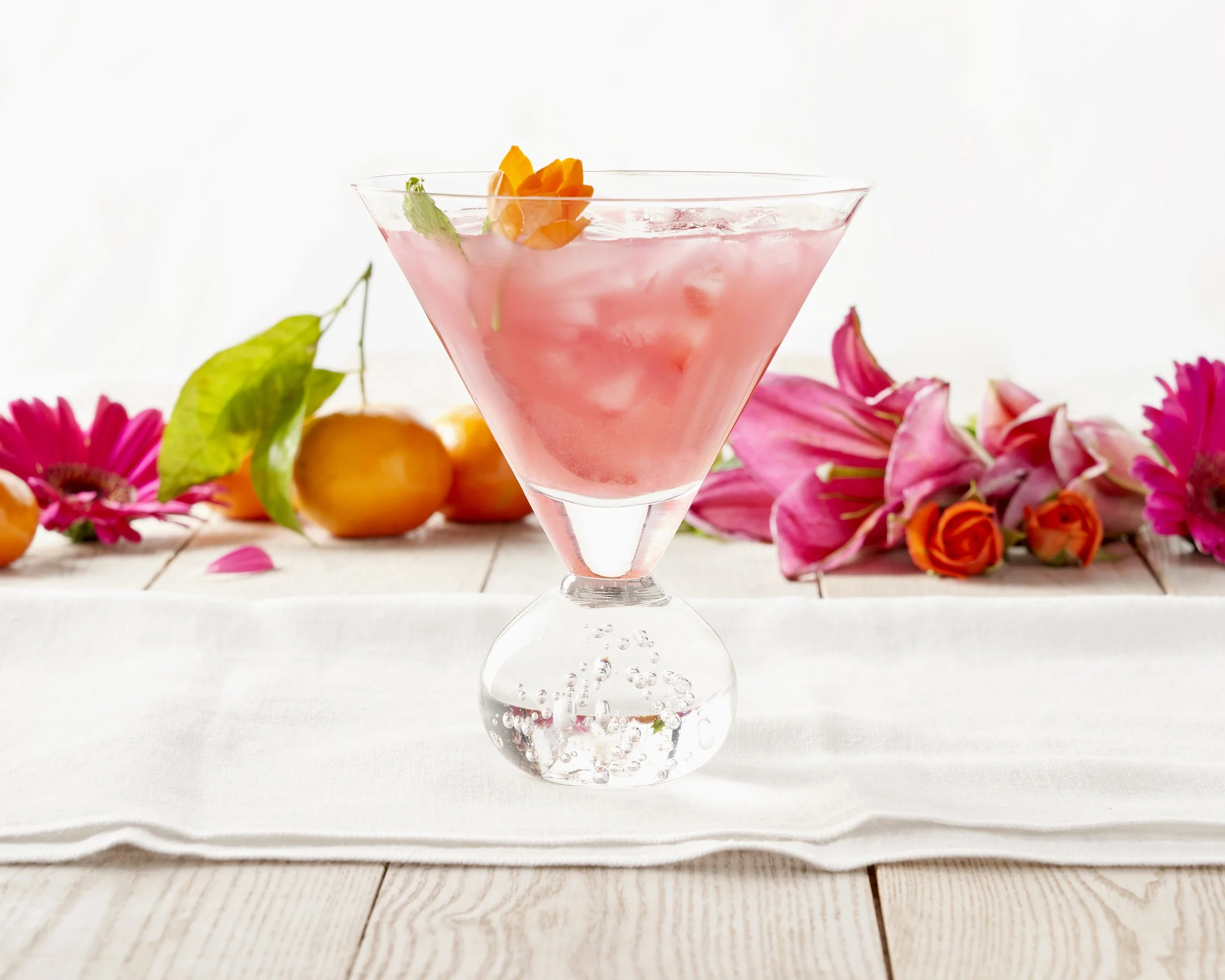 A pink cocktail in a martini glass garnished with an orange flower, on a white tablecloth with colorful flowers and oranges in the background.