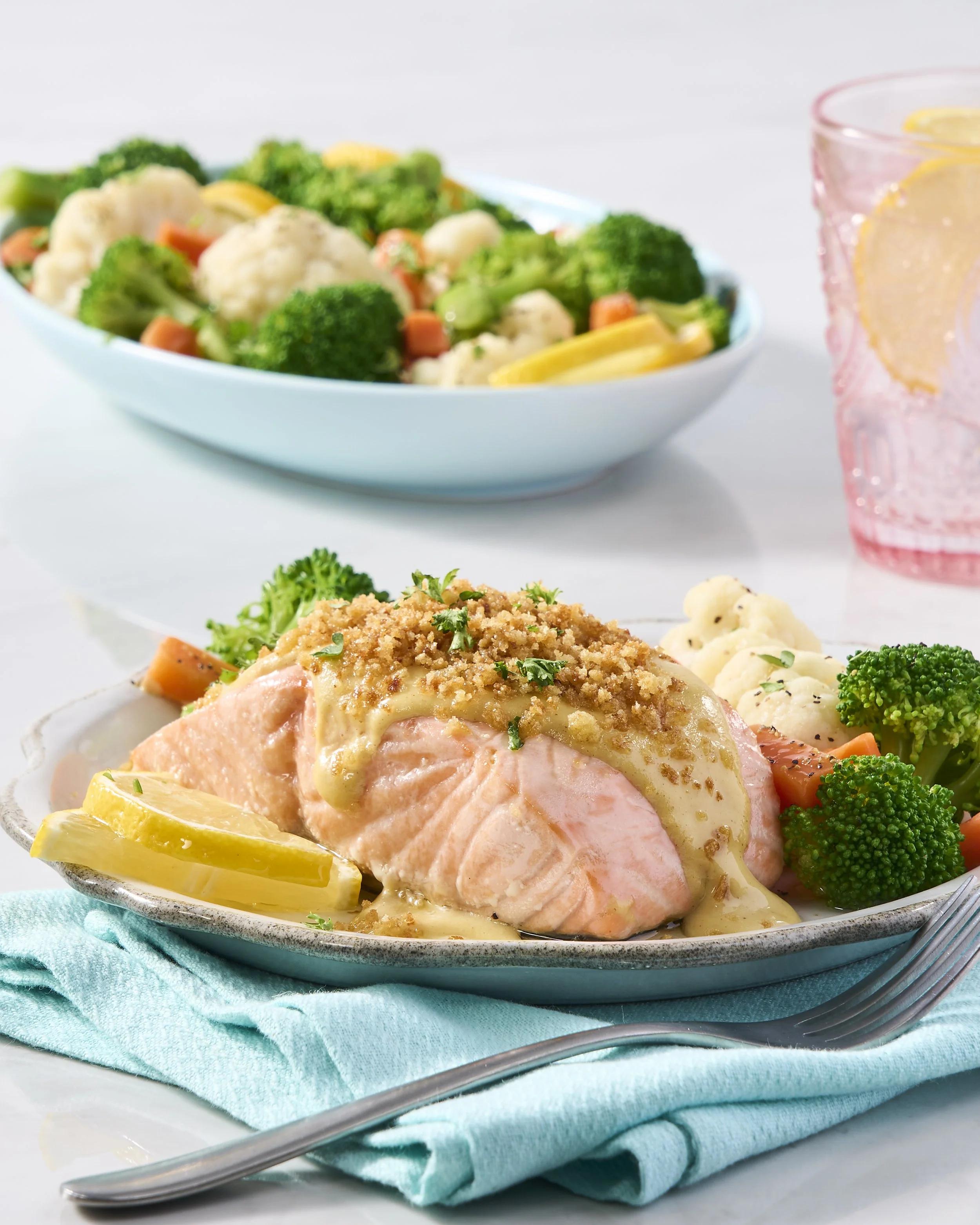 Baked salmon topped with lemon, served with broccoli, cauliflower, and carrots, on a decorative plate with lemon wedges, placed on a blue cloth with a fork nearby.