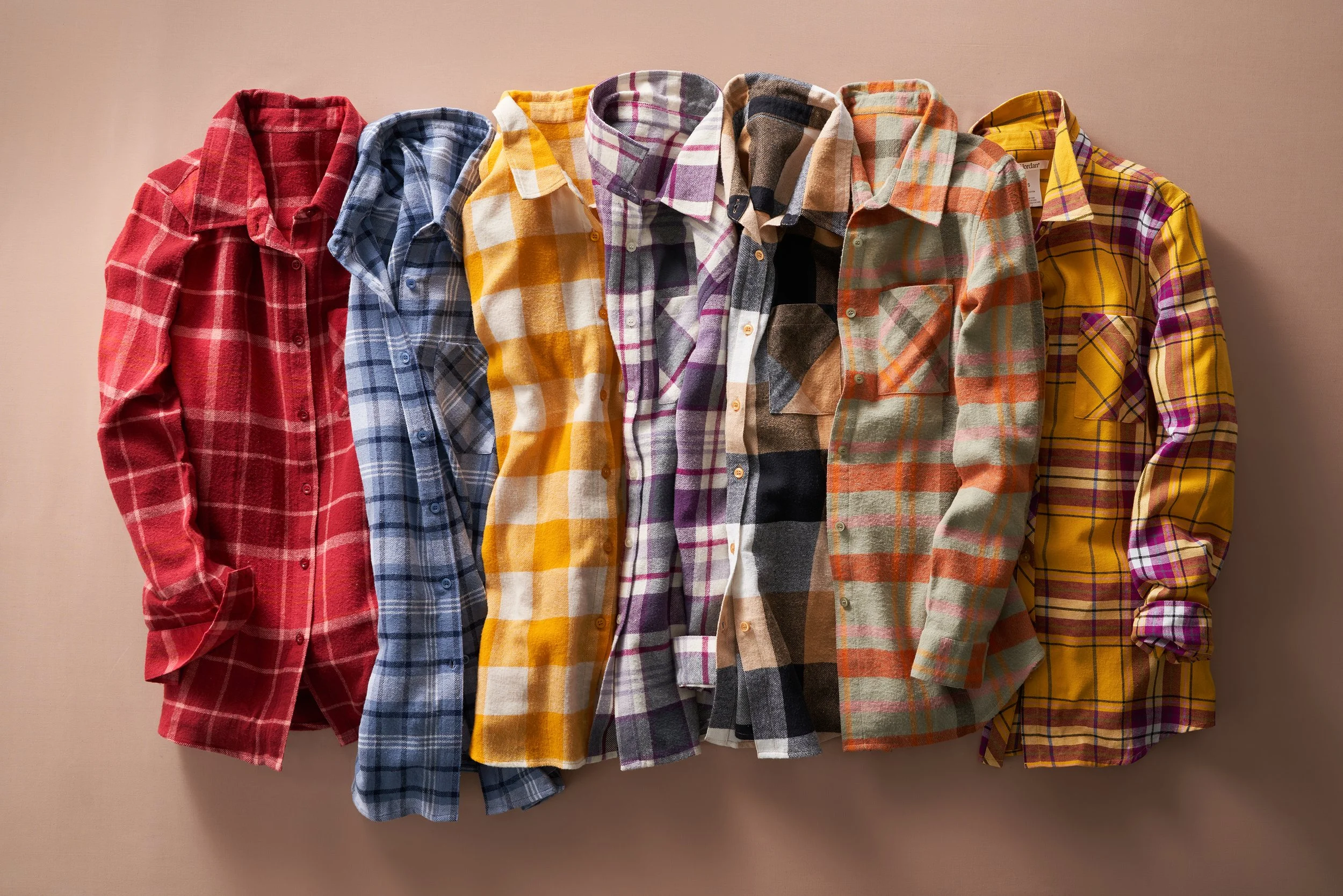 Six plaid flannel shirts in red, blue, yellow, purple, black, and orange hanging on a wall.