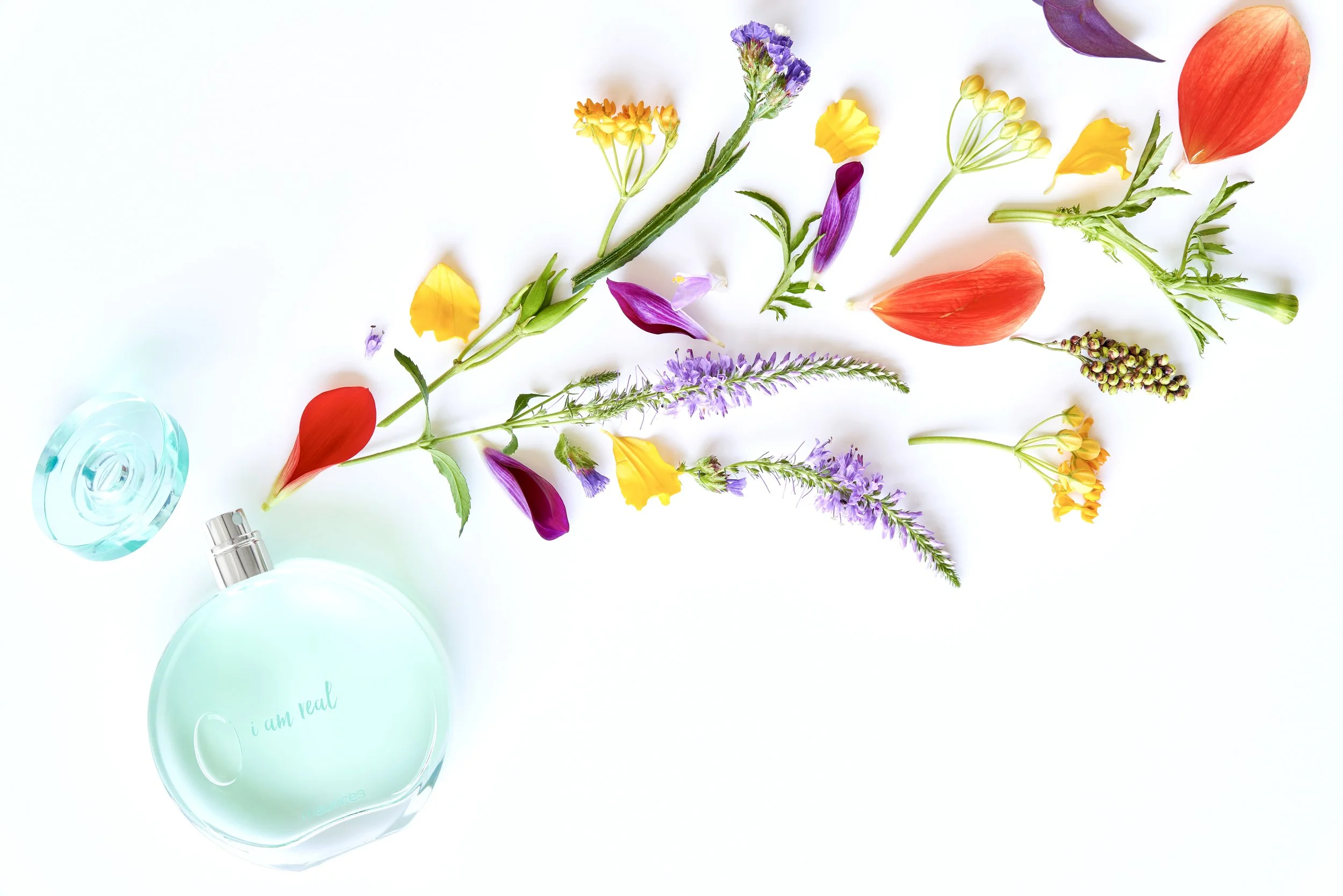 A perfume bottle with the words 'i am real' written on it, surrounded by colorful flower petals and sprigs on a white background.