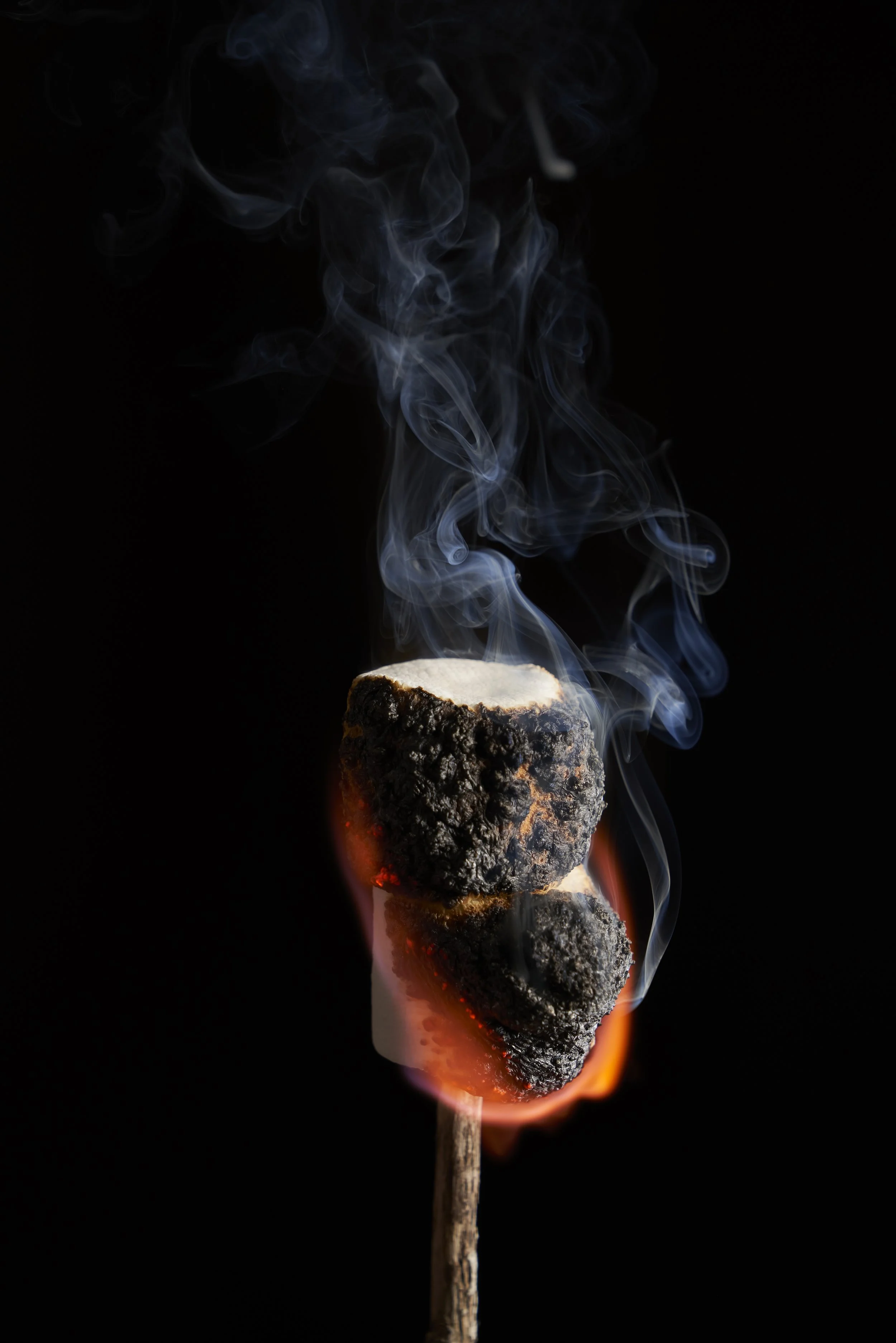 Burning marshmallow on a stick with smoke rising against a black background.
