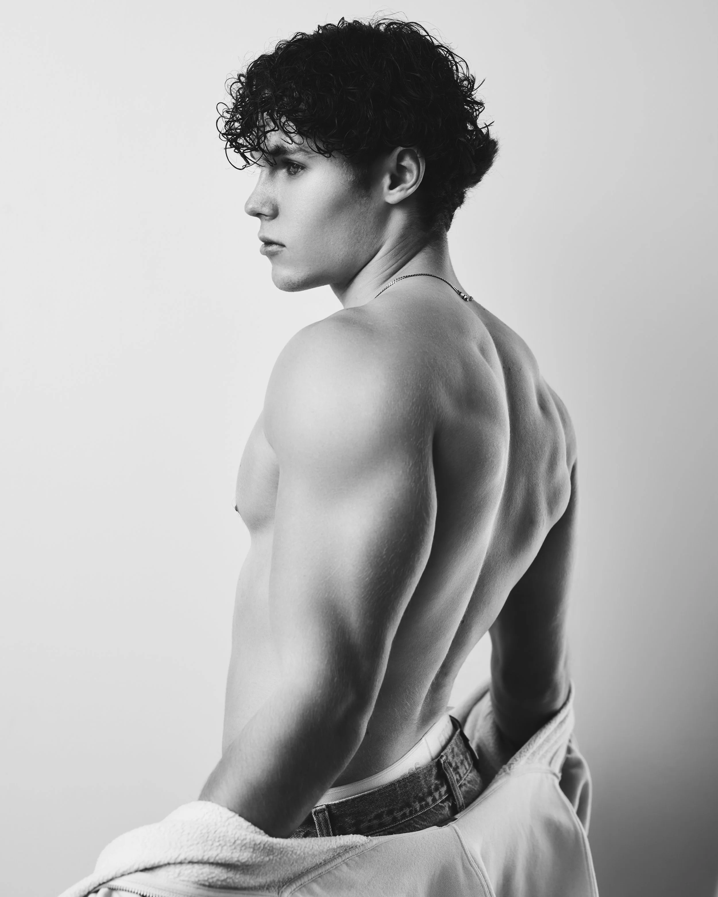 Black and white photo of a young man with curly hair looking to the side, shirtless, with muscular back and arm muscles, holding a jacket.