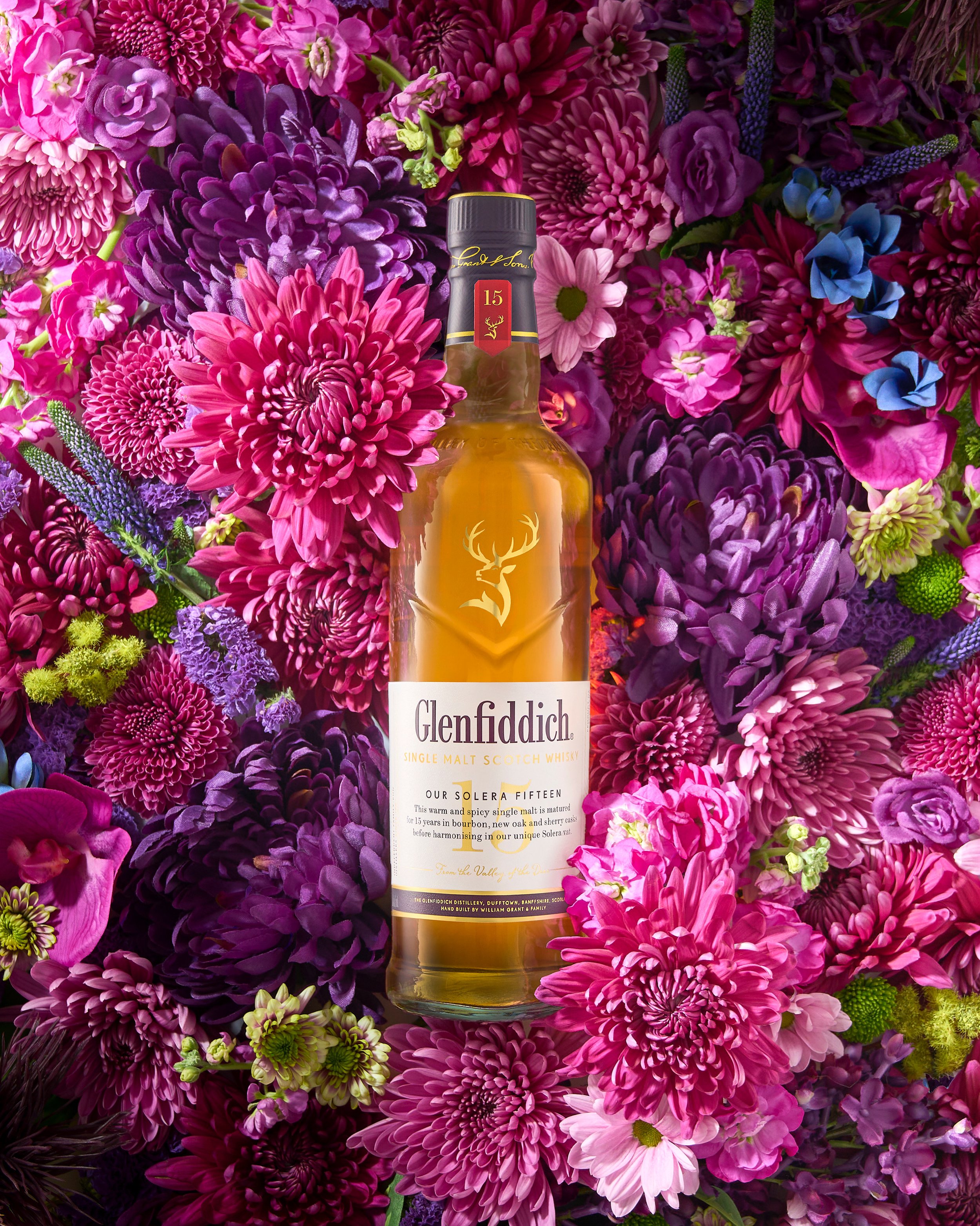 A bottle of Glenfiddich Single Malt Scotch Whisky surrounded by pink, purple, and violet flowers.
