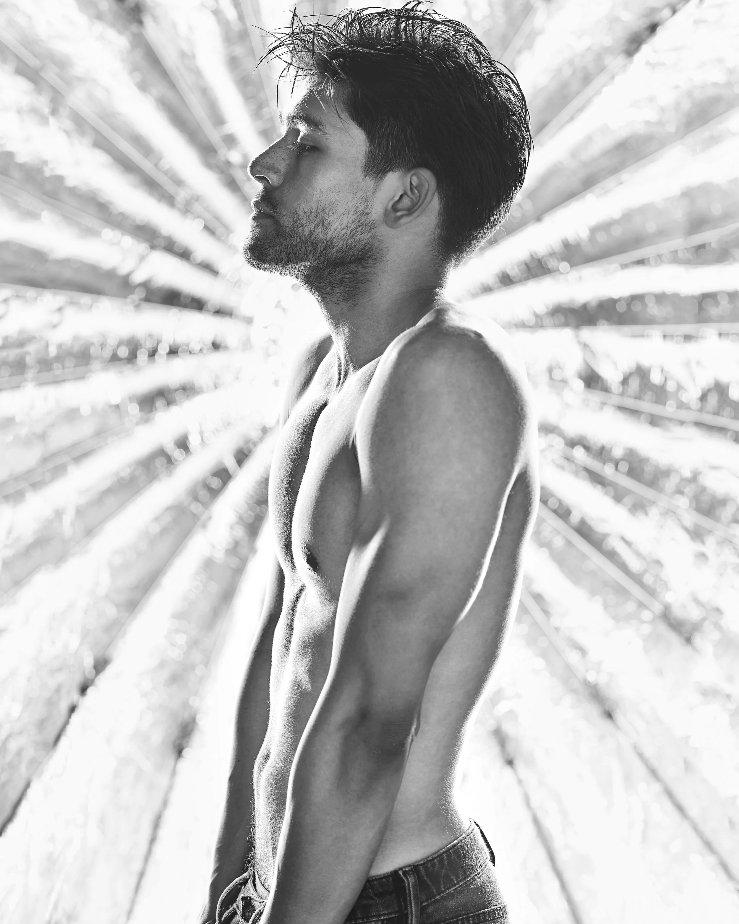 Black and white photo of a shirtless young man with closed eyes and tousled hair, standing in front of a radial pattern of light and shadow.