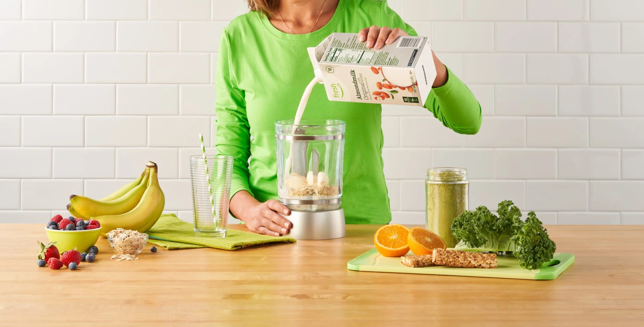 A woman in a bright green shirt making a smoothie in a blender. She is pouring almond milk into the blender. The table has fresh fruits like bananas, strawberries, blueberries, oranges, and kale, along with oats, granola bars, and a jar of powdered s