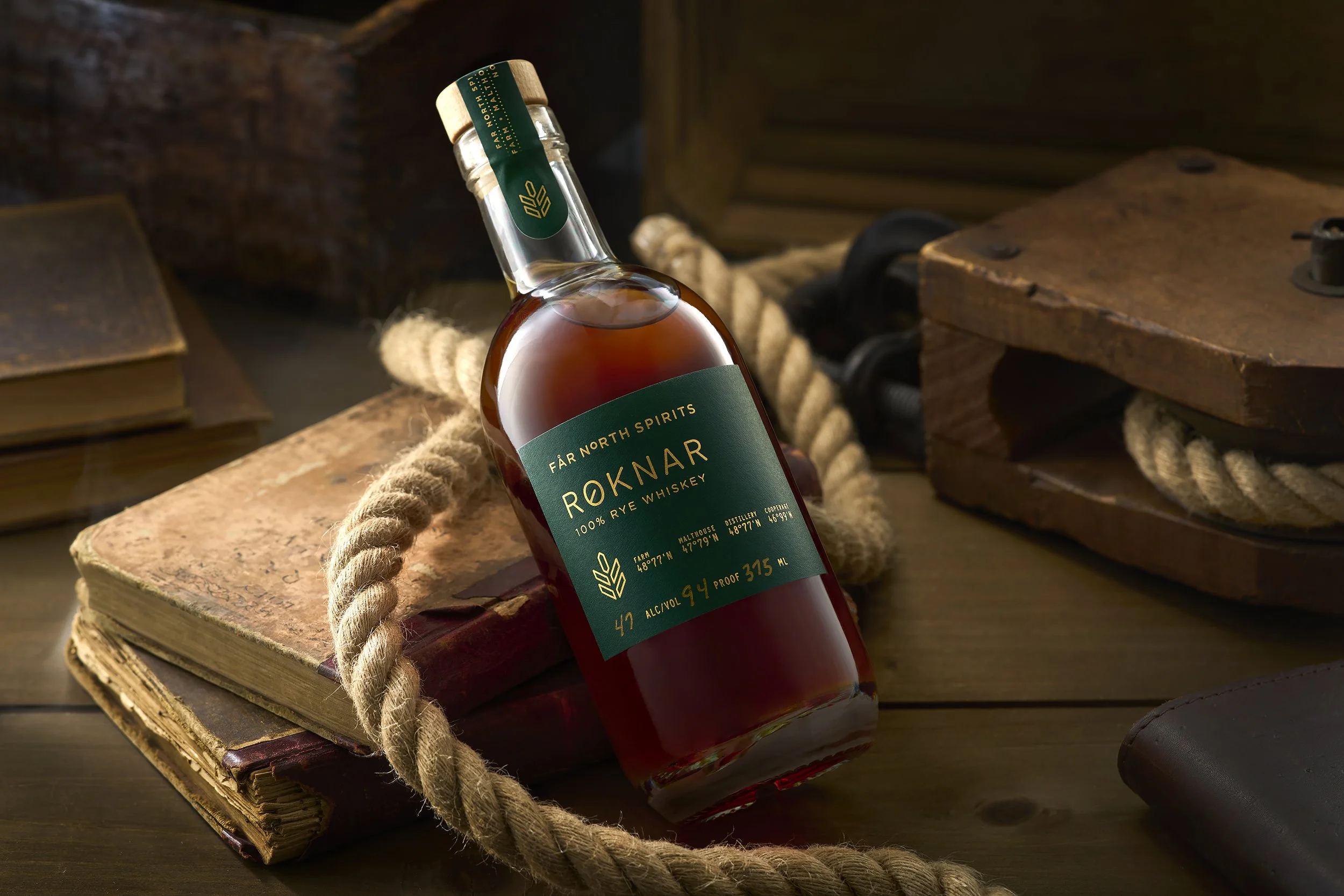 A bottle of Far North Spirits Røknar 100% rye whiskey resting on old stacked books with a thick rope surrounding it, set against a rustic wooden background.