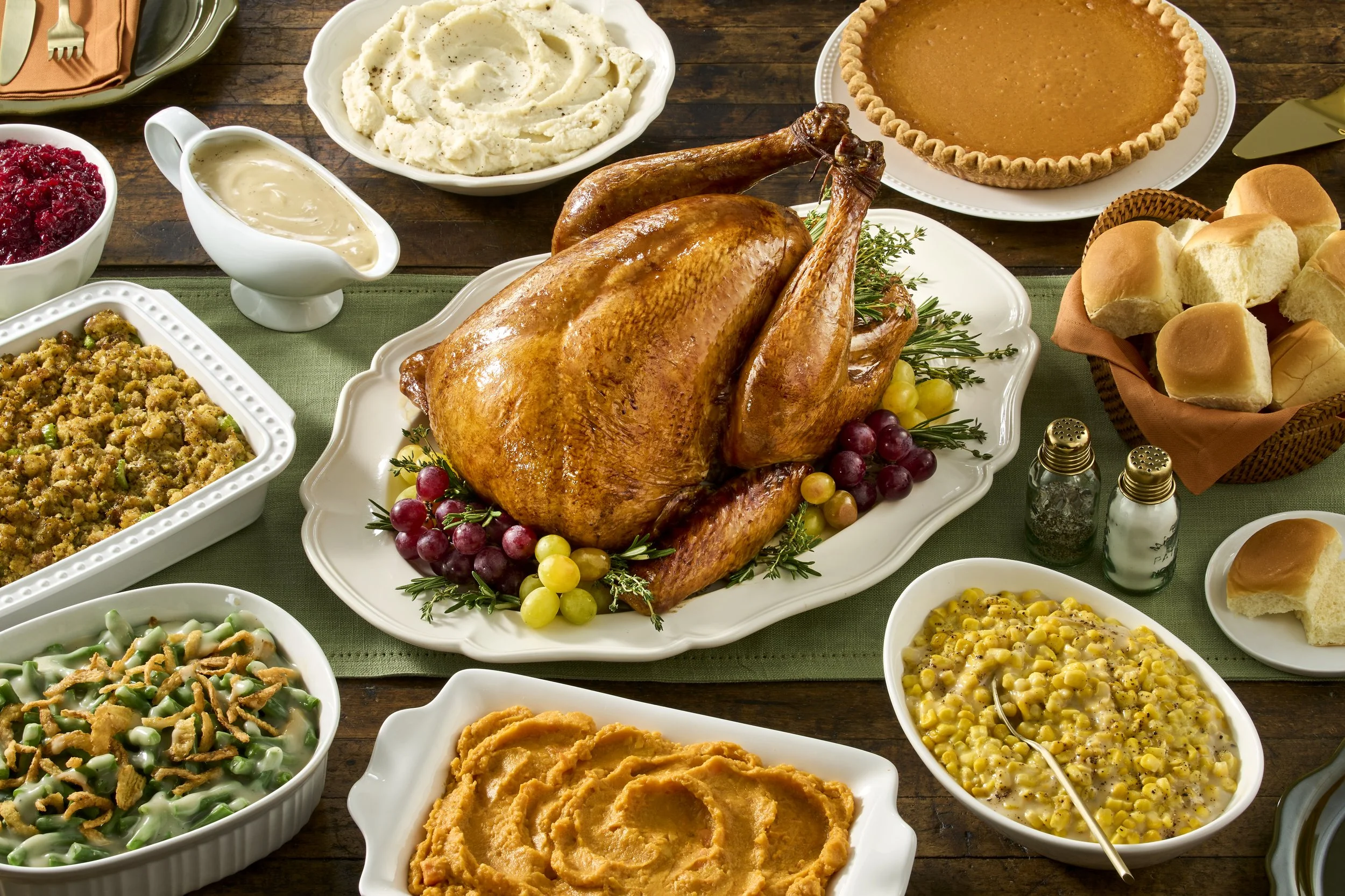 A traditional Thanksgiving feast featuring a roasted turkey with stuffing, mashed potatoes, green bean casserole, sweet potato casserole, cranberry sauce, dinner rolls, pumpkin pie, and other side dishes on a wooden table.