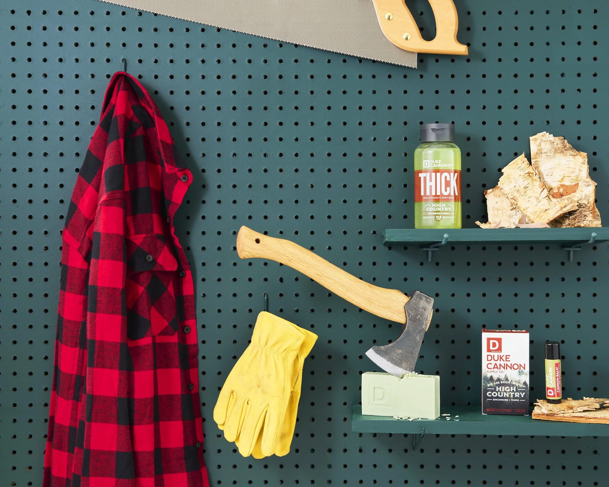 A red and black checkered flannel shirt hanging on a dark teal pegboard wall alongside a pair of yellow work gloves. A hand saw with a silver blade and wooden handle, an axe embedded in soap, a bottle of green thick body wash, chopped firewood, and a