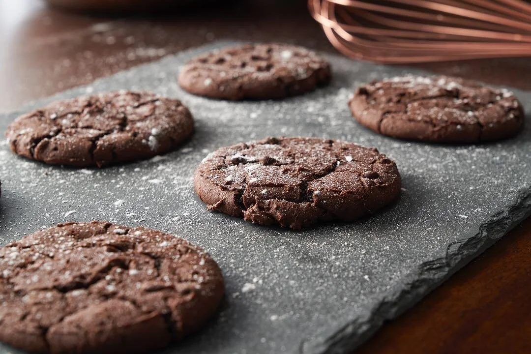 Six chocolate cookies sprinkled with powdered sugar on a black slate serving board.
