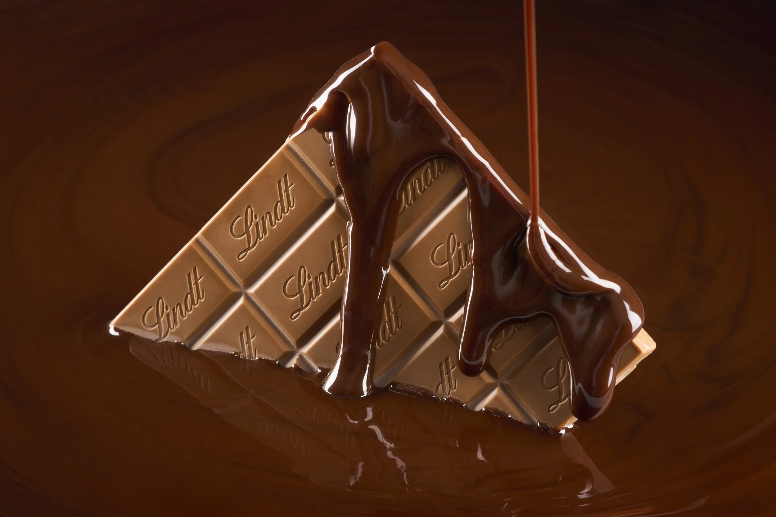 Piece of milk chocolate labeled 'Lindt' being coated with flowing chocolate.