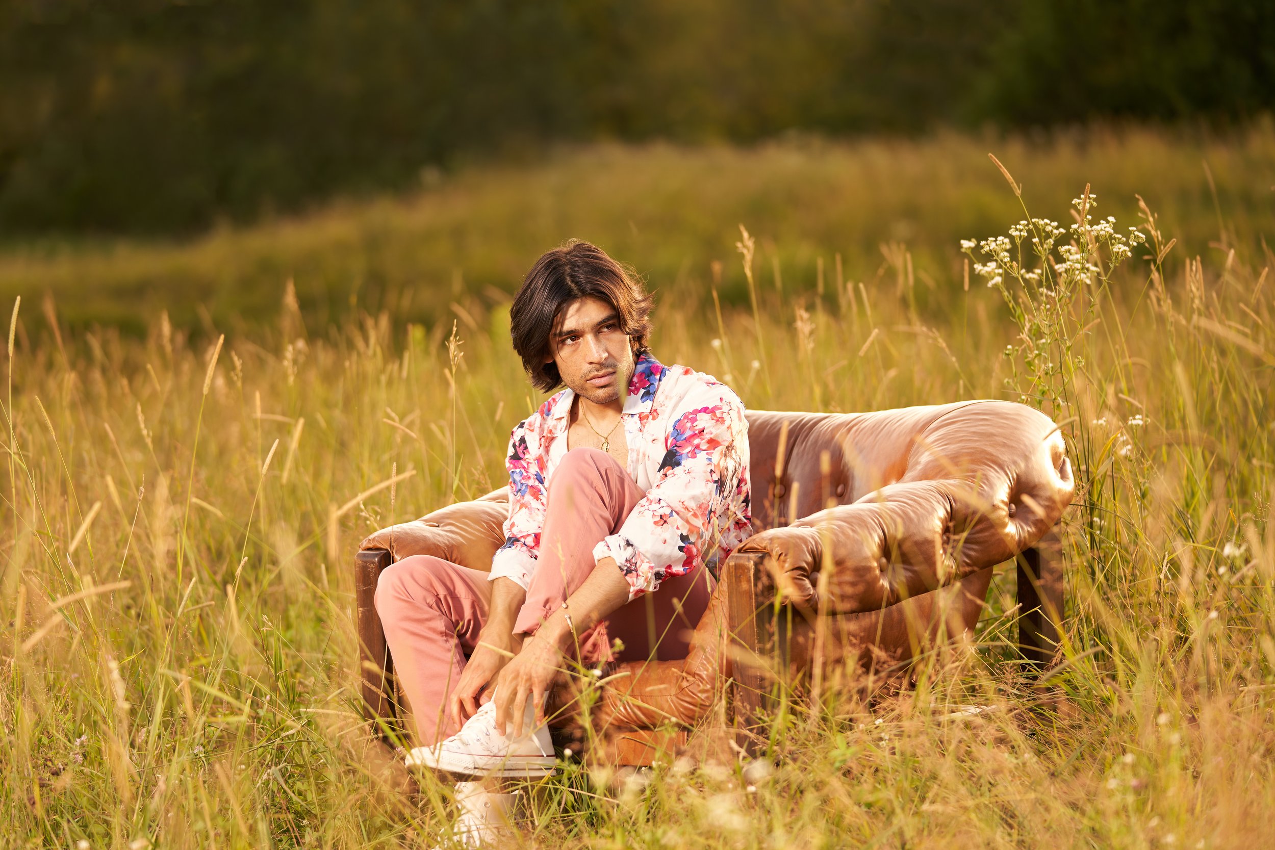 A young man sitting on a vintage brown leather sofa in a grassy field during sunset, wearing a colorful floral shirt and pink pants.