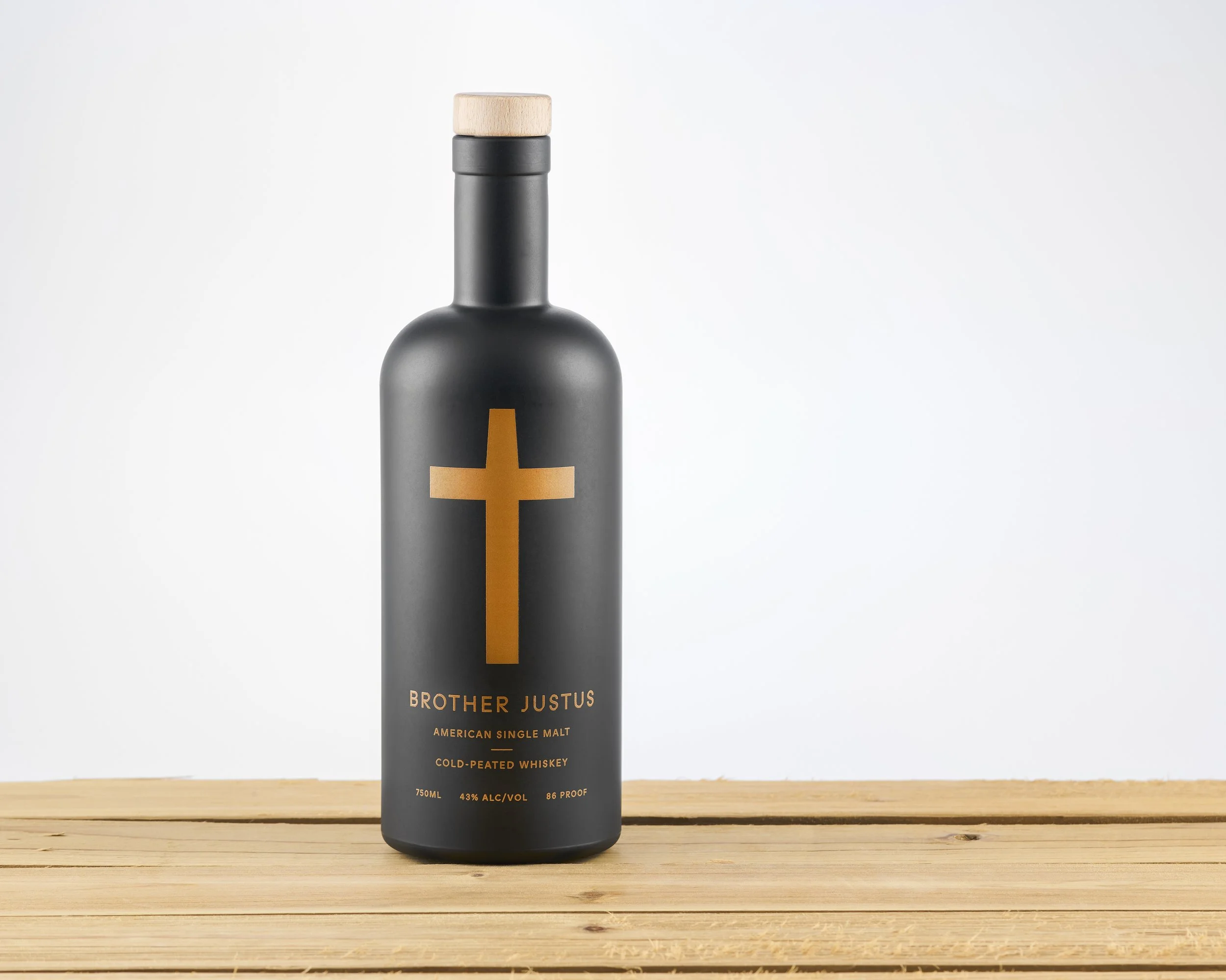 Black bottle with a gold cross label and wooden cap, standing on a wooden surface, and a plain light background.