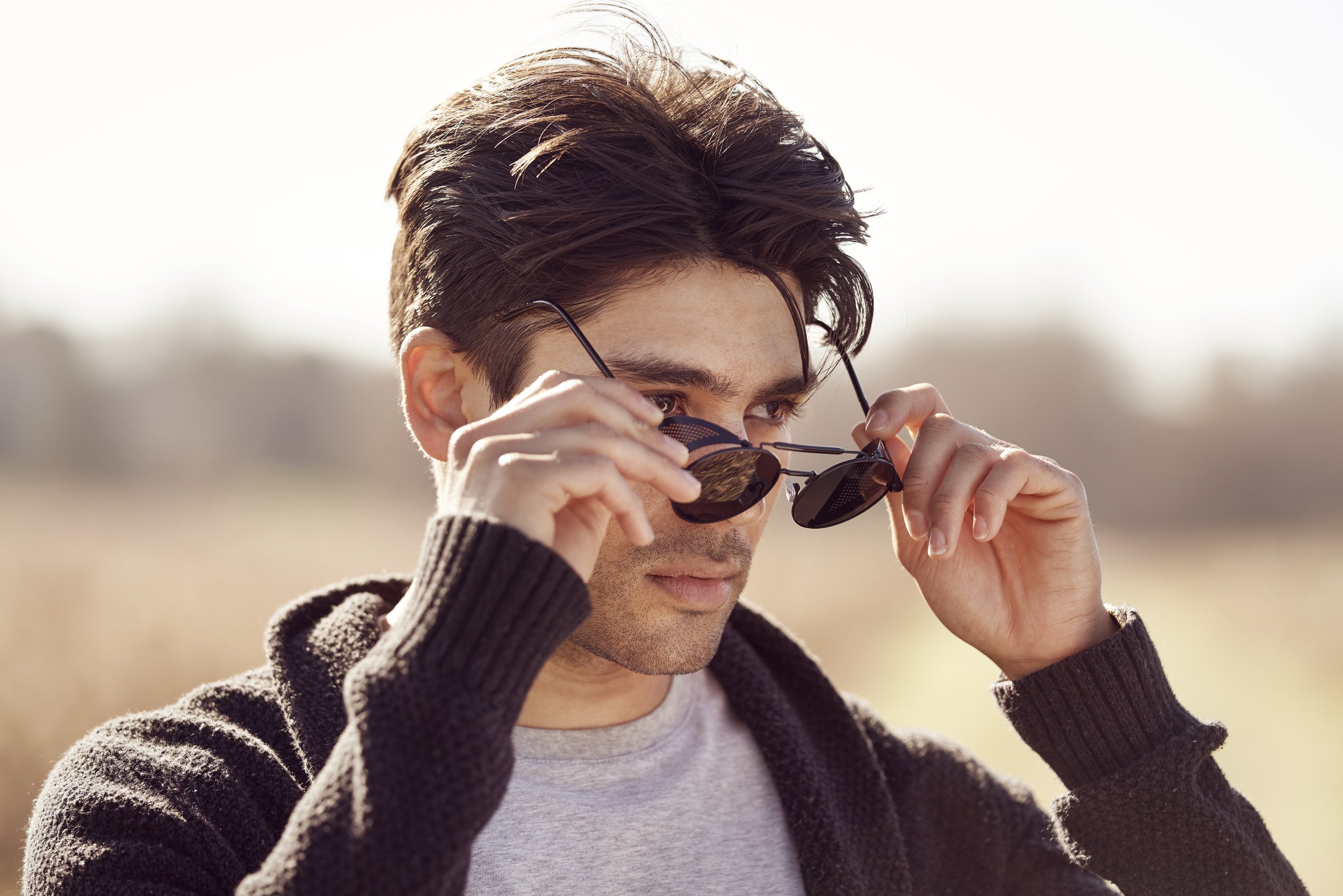 Young man outdoors adjusting his sunglasses, wearing a black hoodie and a light gray t-shirt, with a blurred natural background.