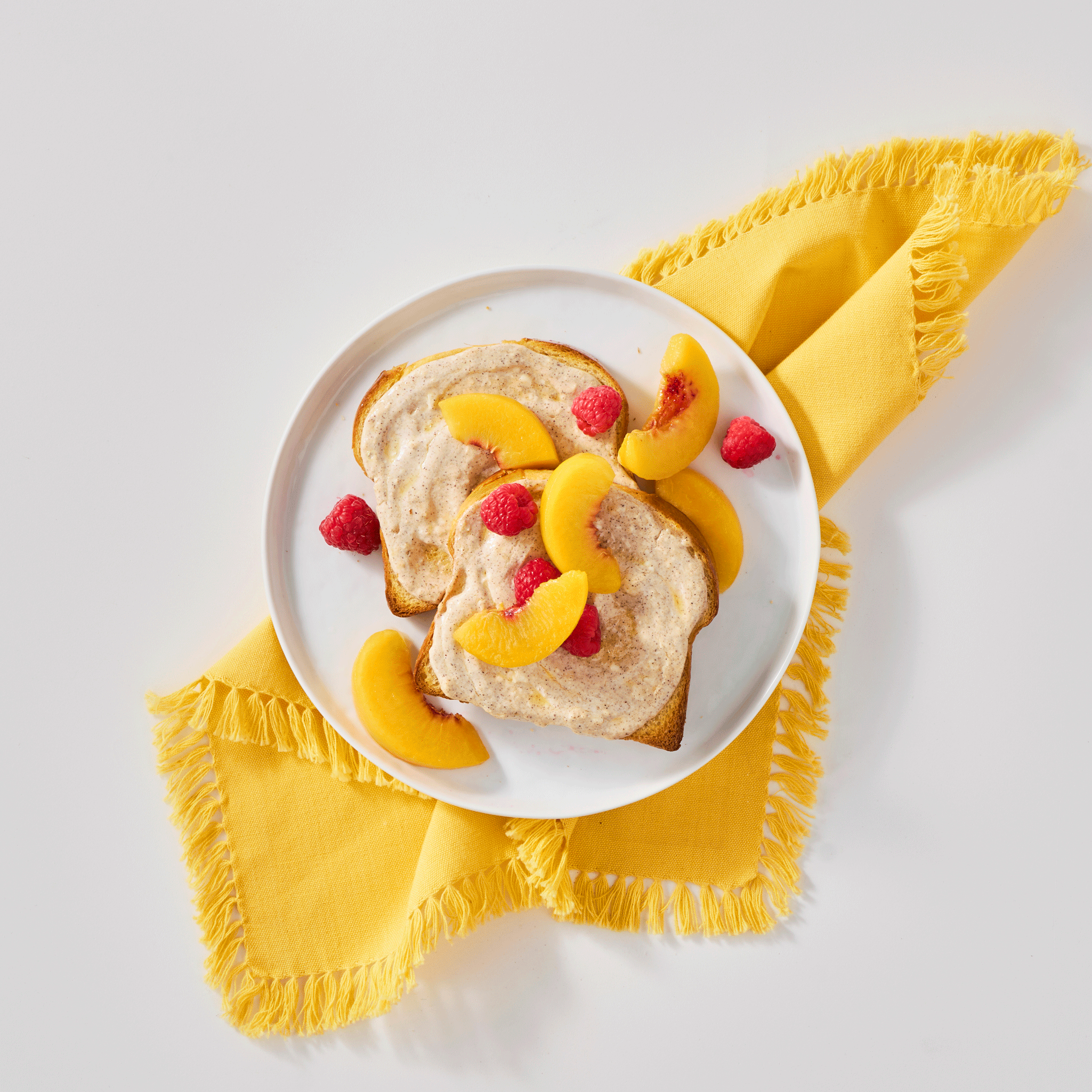 Slices of toast topped with creamy spread, garnished with peach slices and raspberries on a white plate, set on a yellow napkin.